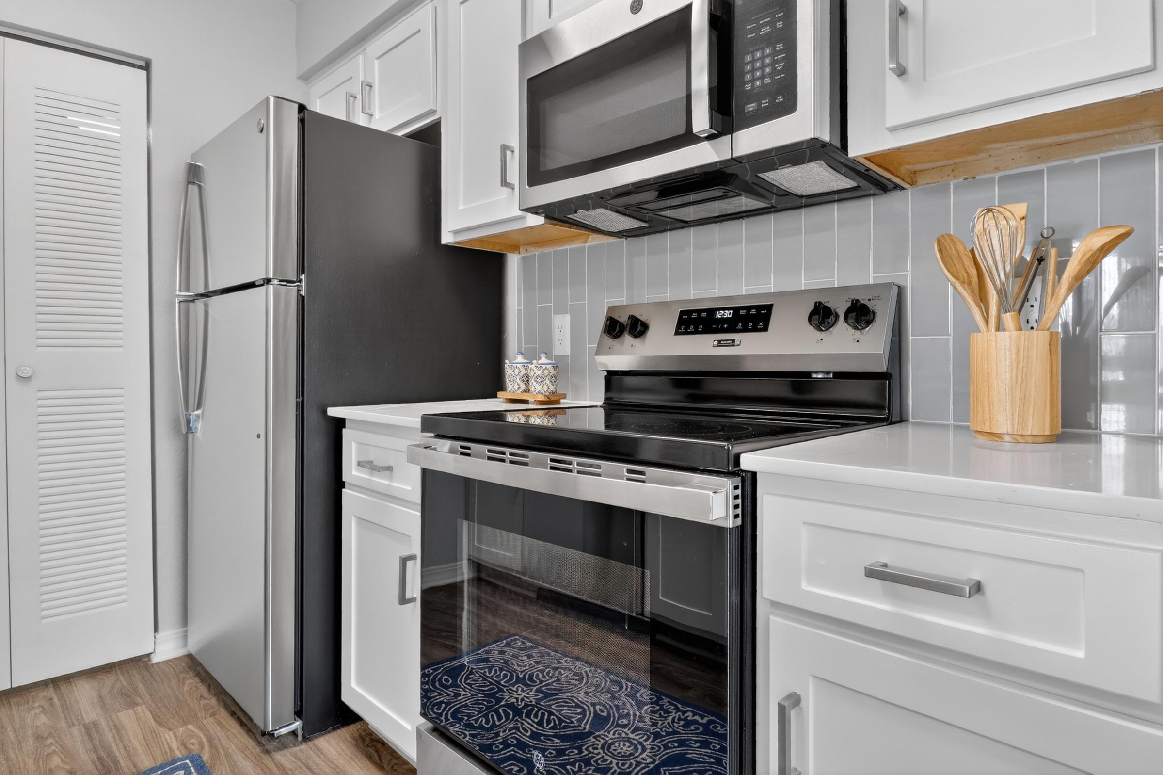 A modern kitchen featuring stainless steel appliances, including a black microwave and stove, with white cabinets and a light gray backsplash. A wooden utensil holder is on the counter, and a decorative rug is visible on the floor. The refrigerator is black, complementing the overall sleek design.