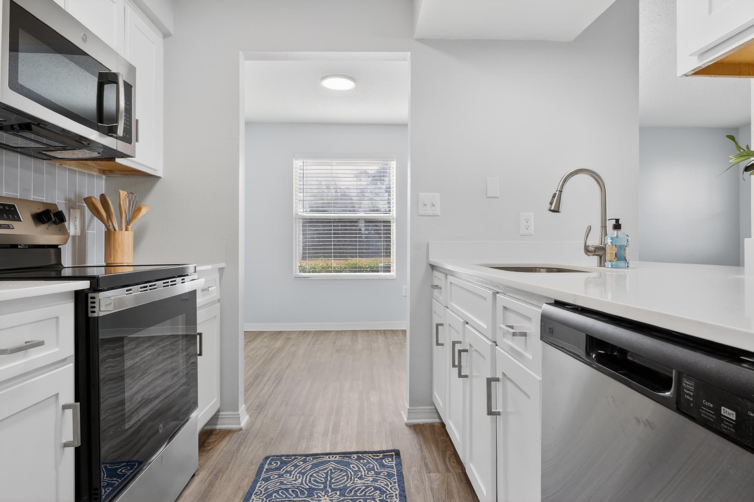 A modern kitchen featuring white cabinets, stainless steel appliances, and a quartz countertop. The view opens into a bright room with a window and light-colored walls. A blue decorative rug is placed on the floor in front of the sink, enhancing the clean and contemporary aesthetic.