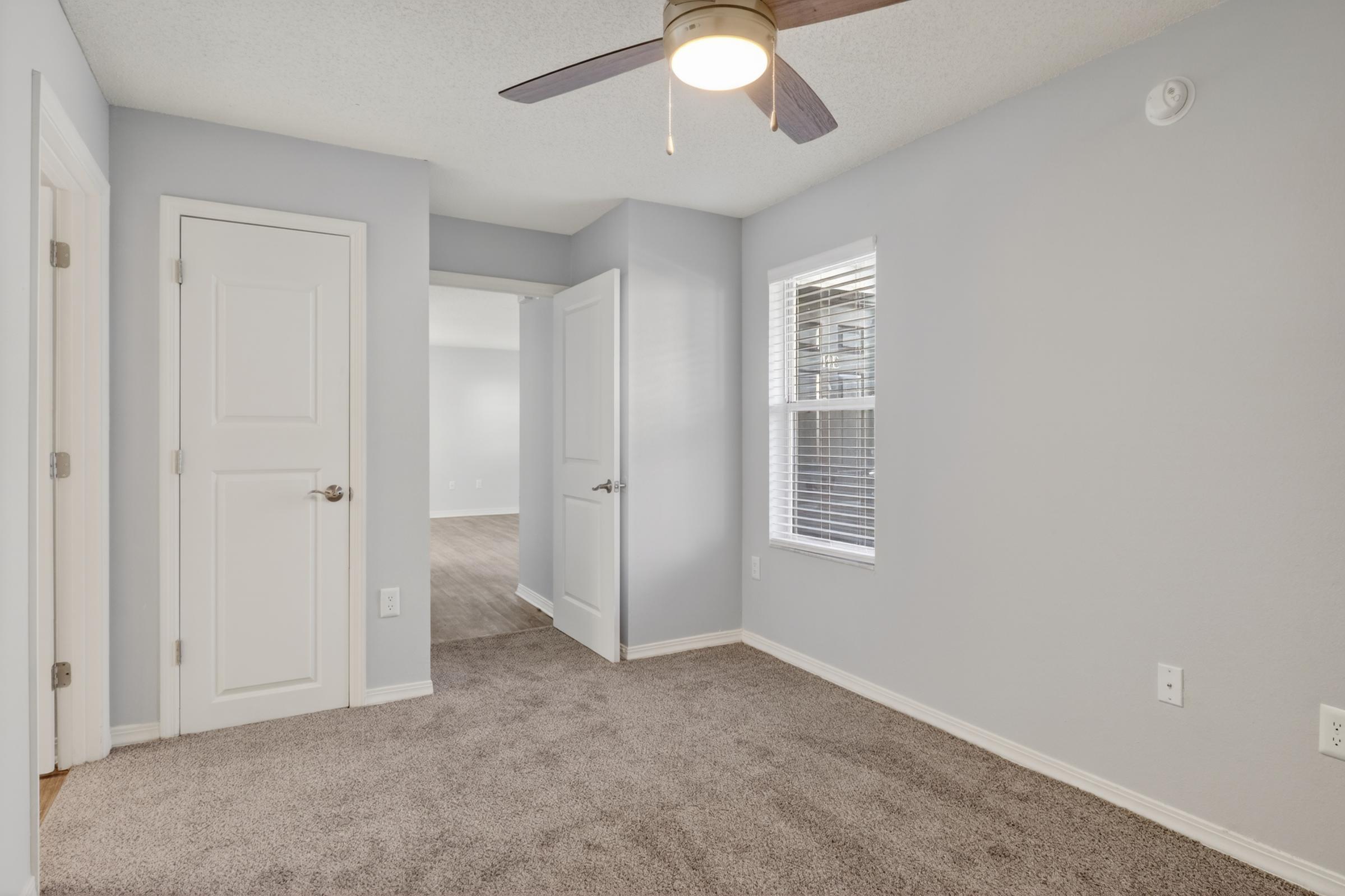 Empty room featuring light gray walls and carpeted flooring, with a ceiling fan and a window providing natural light. The image shows two doorways leading to adjacent rooms. The overall layout appears spacious and well-lit, with neutral decor.