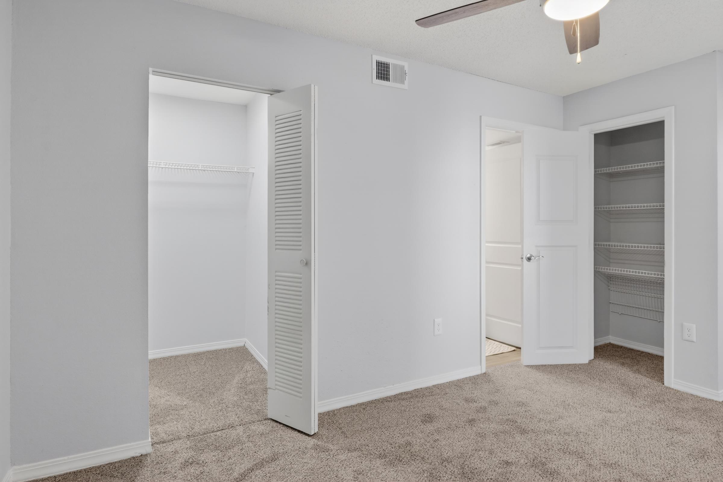 A well-lit, modern bedroom featuring light gray walls and carpeted flooring. Two doors are visible: one leading to a small closet and the other to a bathroom or additional storage area. A ceiling fan hangs from the ceiling, adding to the room's comfort and appeal.