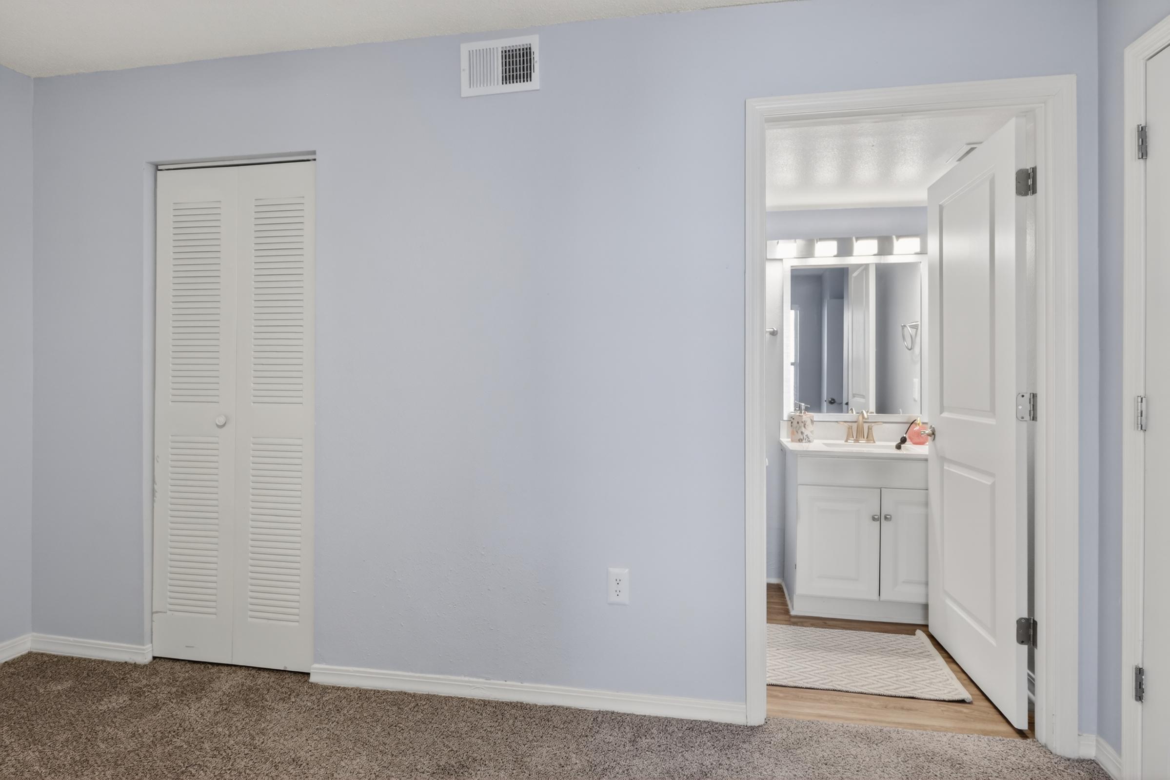 A light blue room featuring a closed white closet door on the left and an open door leading to a bathroom on the right. The bathroom has a mirror with lights above the sink, and a light rug on the floor. The carpeted floor adds warmth to the space.