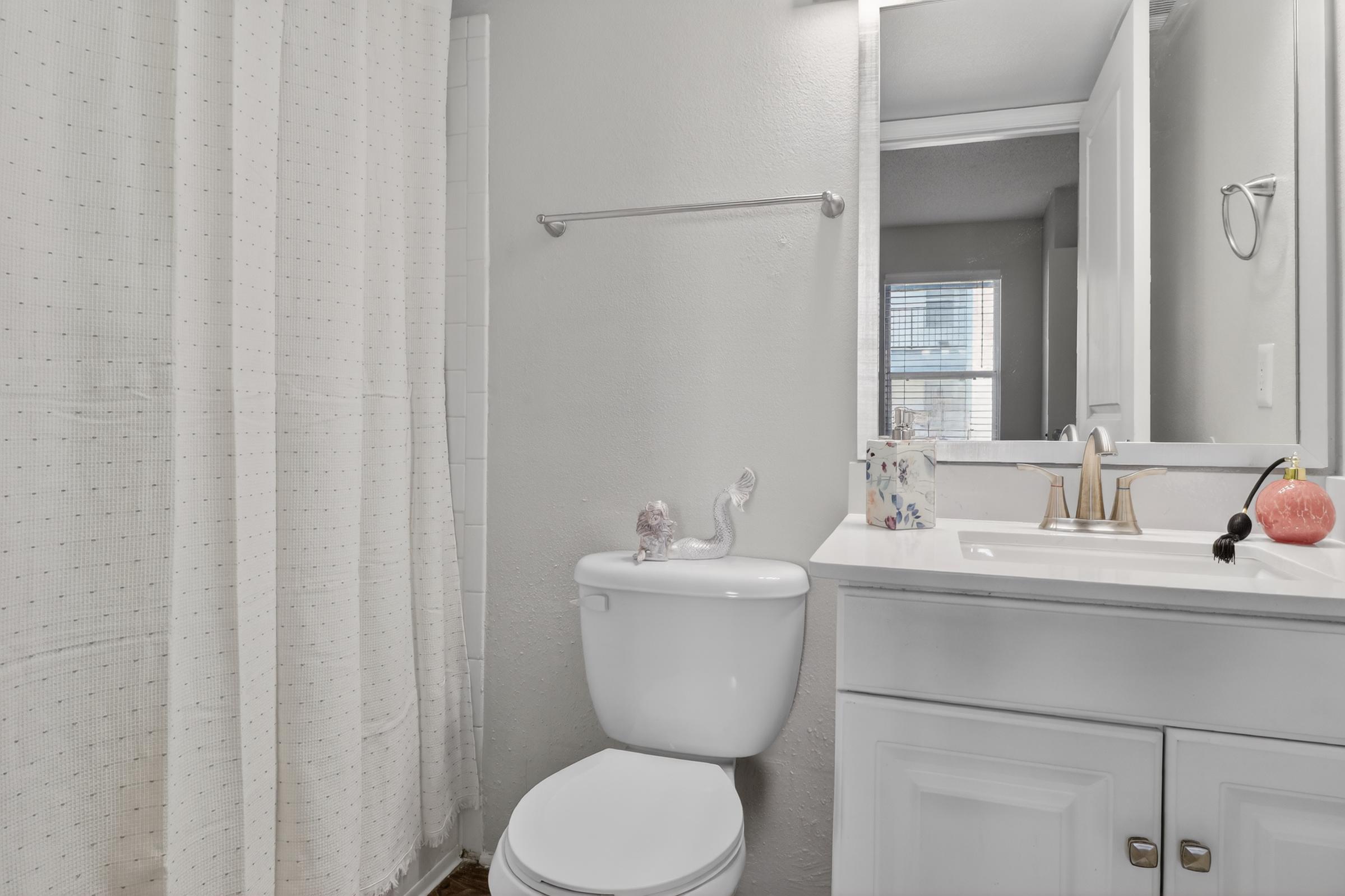A clean bathroom featuring a white toilet, a mirror above a sink with gold faucets, and a shower curtain. There is a decorative item on the sink and natural light entering through a window. The walls are light-colored, creating a bright, airy atmosphere.
