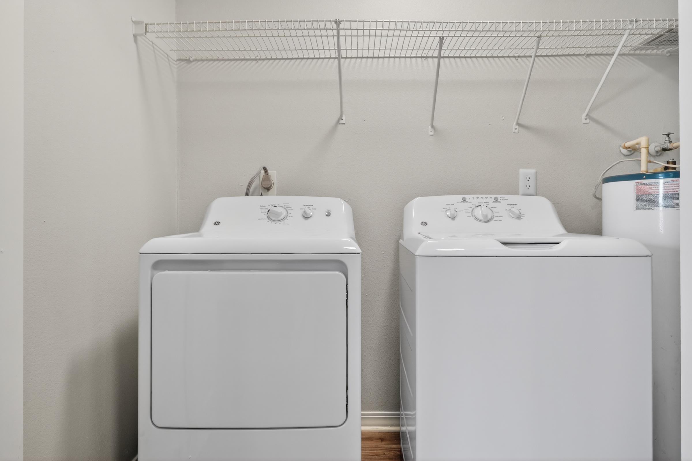 A clean, organized laundry room featuring a white washing machine and dryer side by side. Above the machines is a wire shelf for storage, and a water heater is visible to the right against the wall. The walls are a light neutral color, and the floor is wooden.