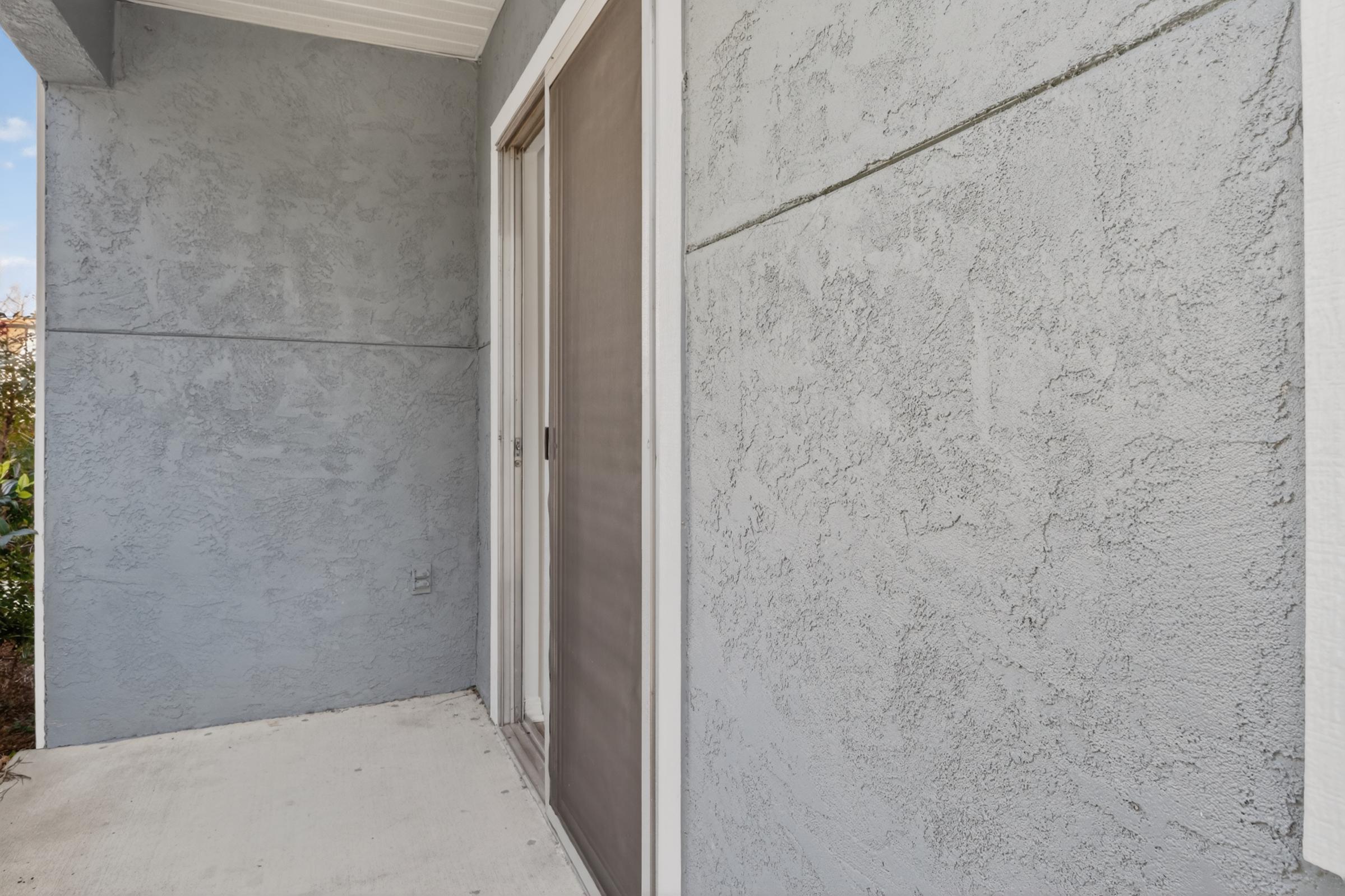 A gray-textured exterior wall with a partially open sliding door and a small porch area. The floor is mostly bare concrete, and there are hints of greenery visible in the background. The scene appears well-lit and offers a view of the surrounding area.