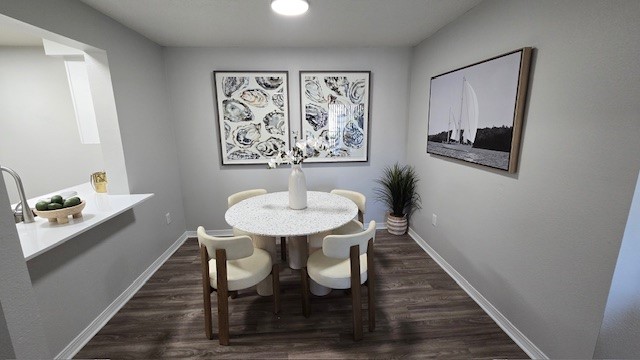 A well-lit dining area featuring a round table with four beige chairs. On the table is a vase with flowers and a fruit bowl. The walls are painted light gray, adorned with two framed artworks. A corner plant adds a touch of greenery, and a small kitchen area is visible in the background.