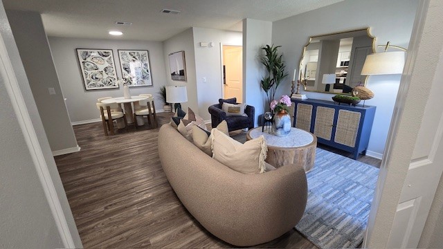 A modern living space featuring a curved beige sofa with decorative pillows, a round wooden coffee table, and an area rug. To the left, a dining area with a circular table and four chairs. A tall plant and a large mirror add elegance, while light walls and wooden flooring create a warm atmosphere.