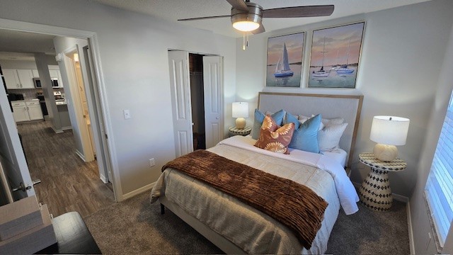 A cozy bedroom featuring a queen-sized bed with decorative pillows, a light brown throw blanket, and two bedside lamps. The walls are adorned with nautical-themed artwork. A ceiling fan hangs above, and there is a carpeted floor. An open doorway leads to a well-lit kitchen area in the background.