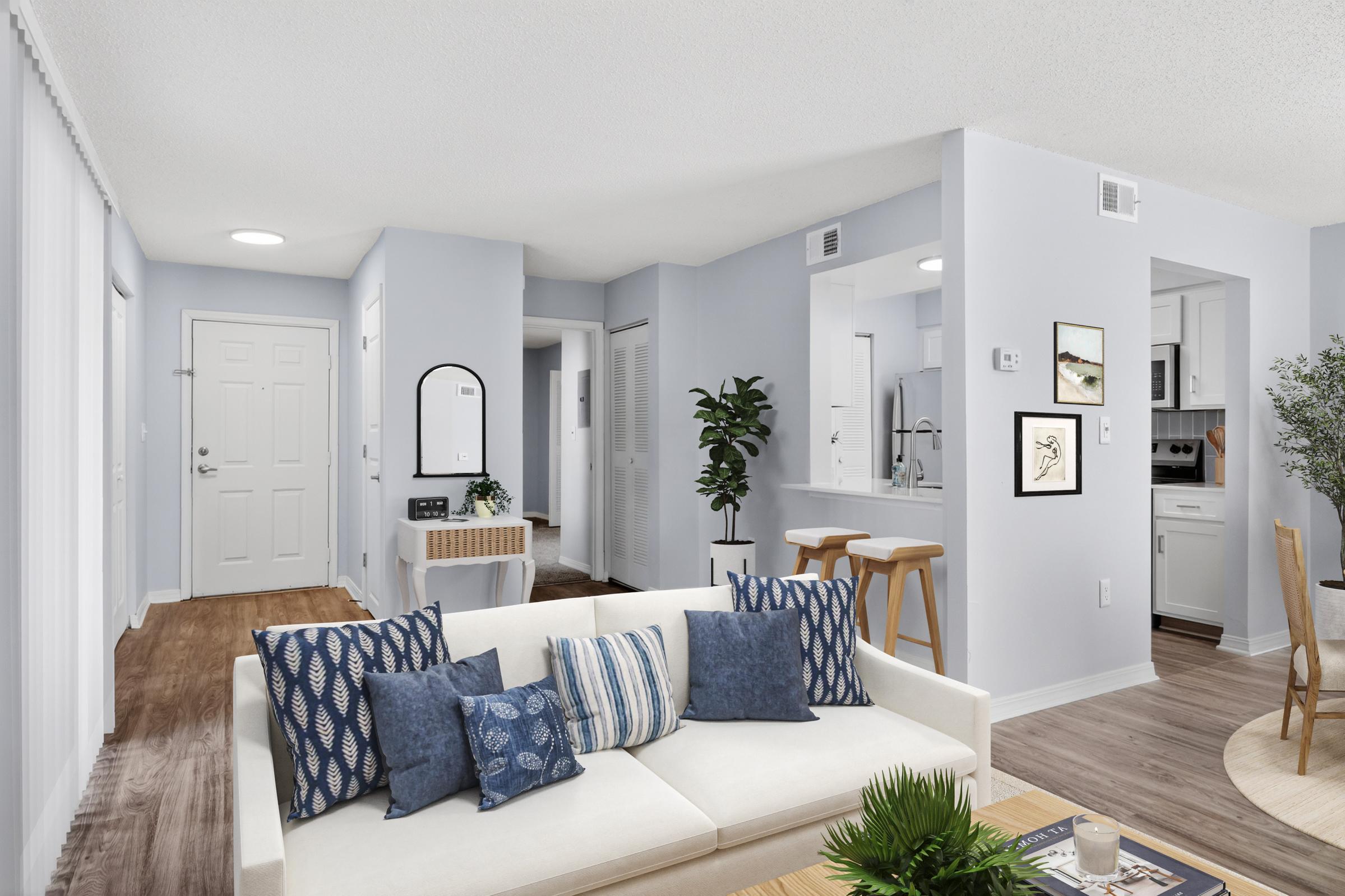A modern, well-lit living space featuring a light-colored sofa with blue accent pillows. There's a small dining area with bar stools, a stylish mirror on the wall, and a plant. The kitchen is partially visible, displaying white cabinetry and a neutral color scheme, creating a welcoming atmosphere.