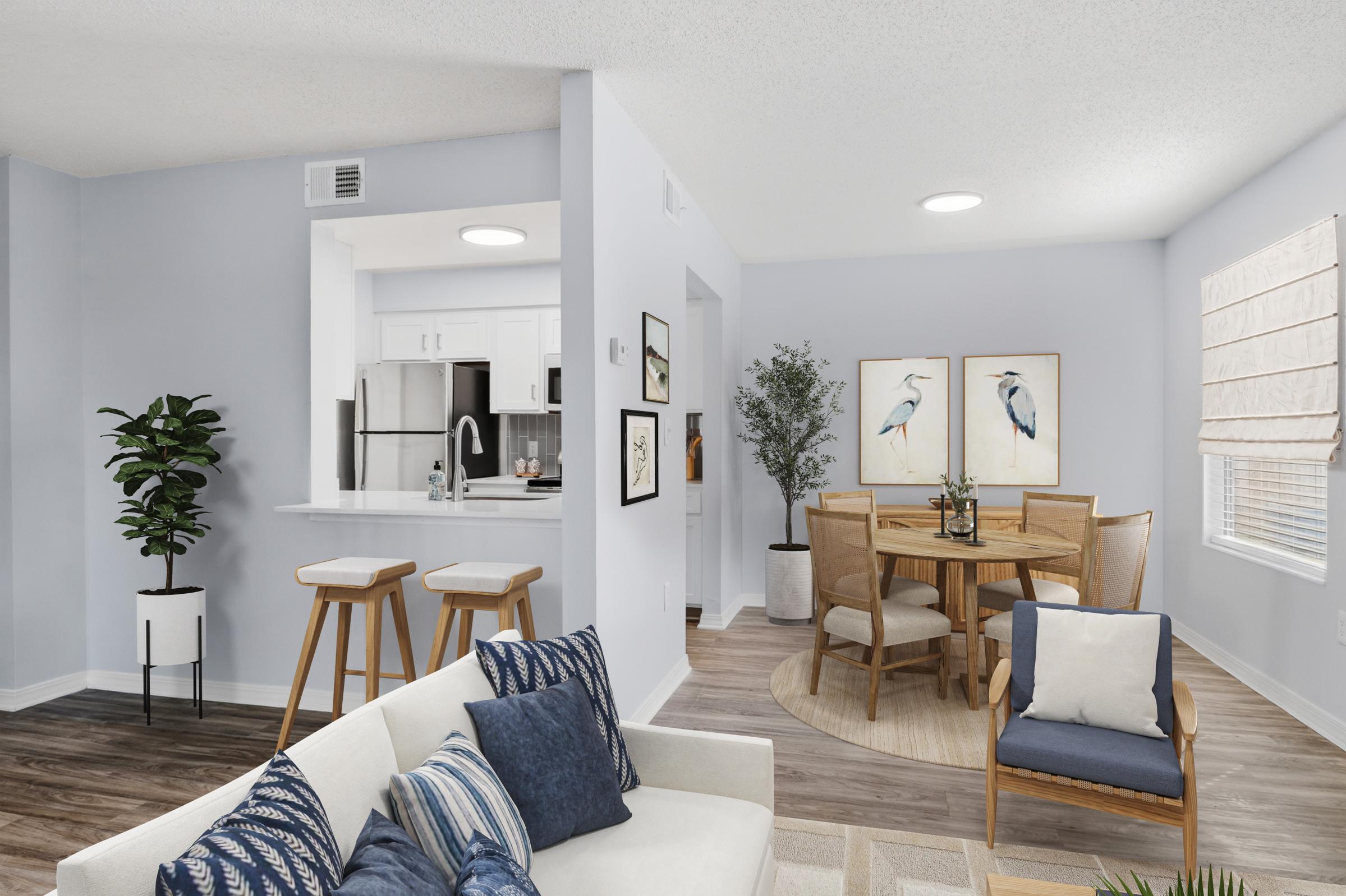 Modern living room and dining area featuring a light blue color scheme. A cozy sofa with decorative pillows, a wooden dining table surrounded by chairs, and a small kitchen area visible in the background. Decor includes potted plants and artwork depicting birds. Natural light enhances the inviting atmosphere.
