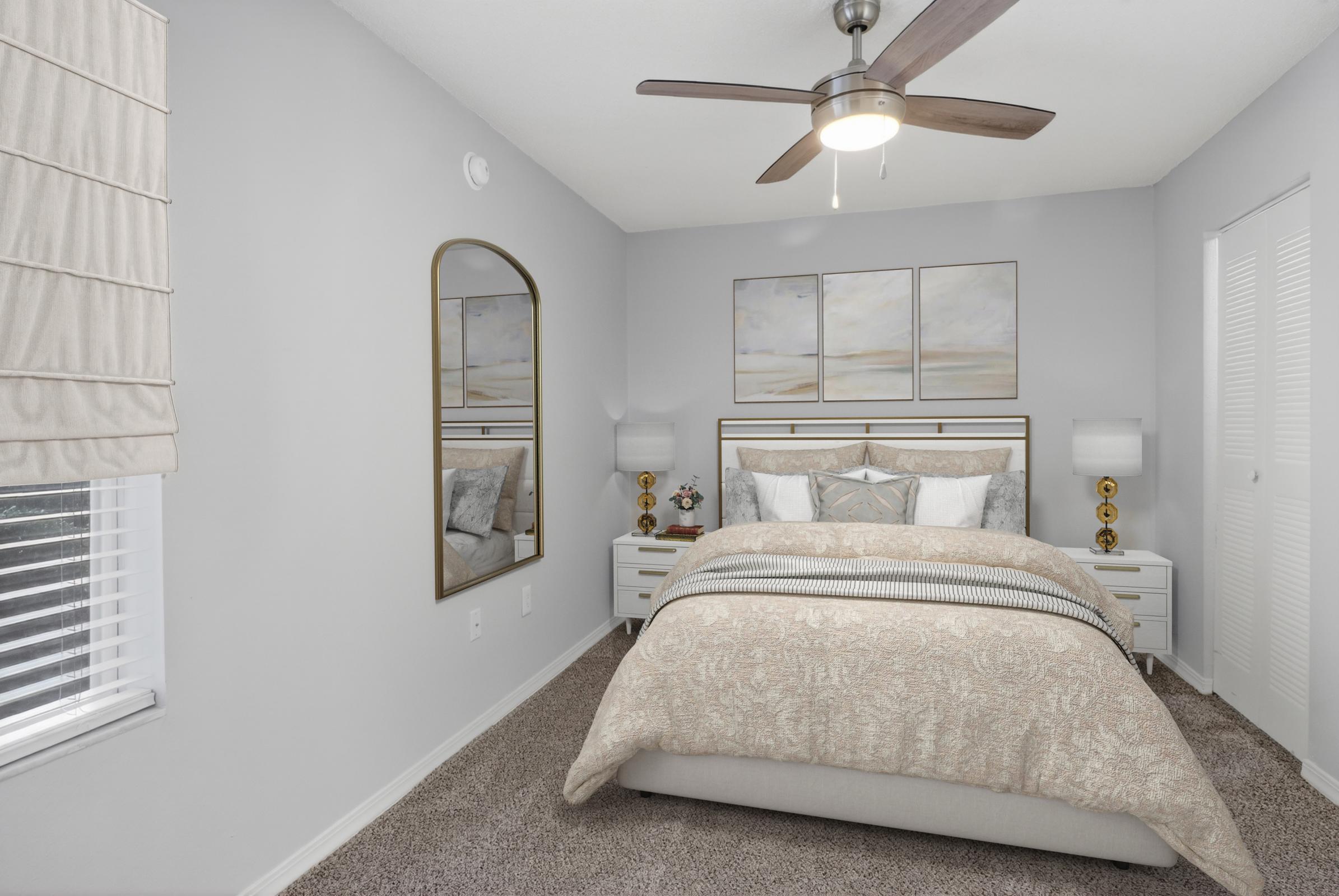 Cozy bedroom featuring a plush bed with light-colored bedding, a stylish mirror, two bedside lamps, and neutral decor. The walls are painted gray, and there's a light-colored area rug on the carpeted floor, creating a warm and inviting atmosphere. A ceiling fan adds modern comfort.