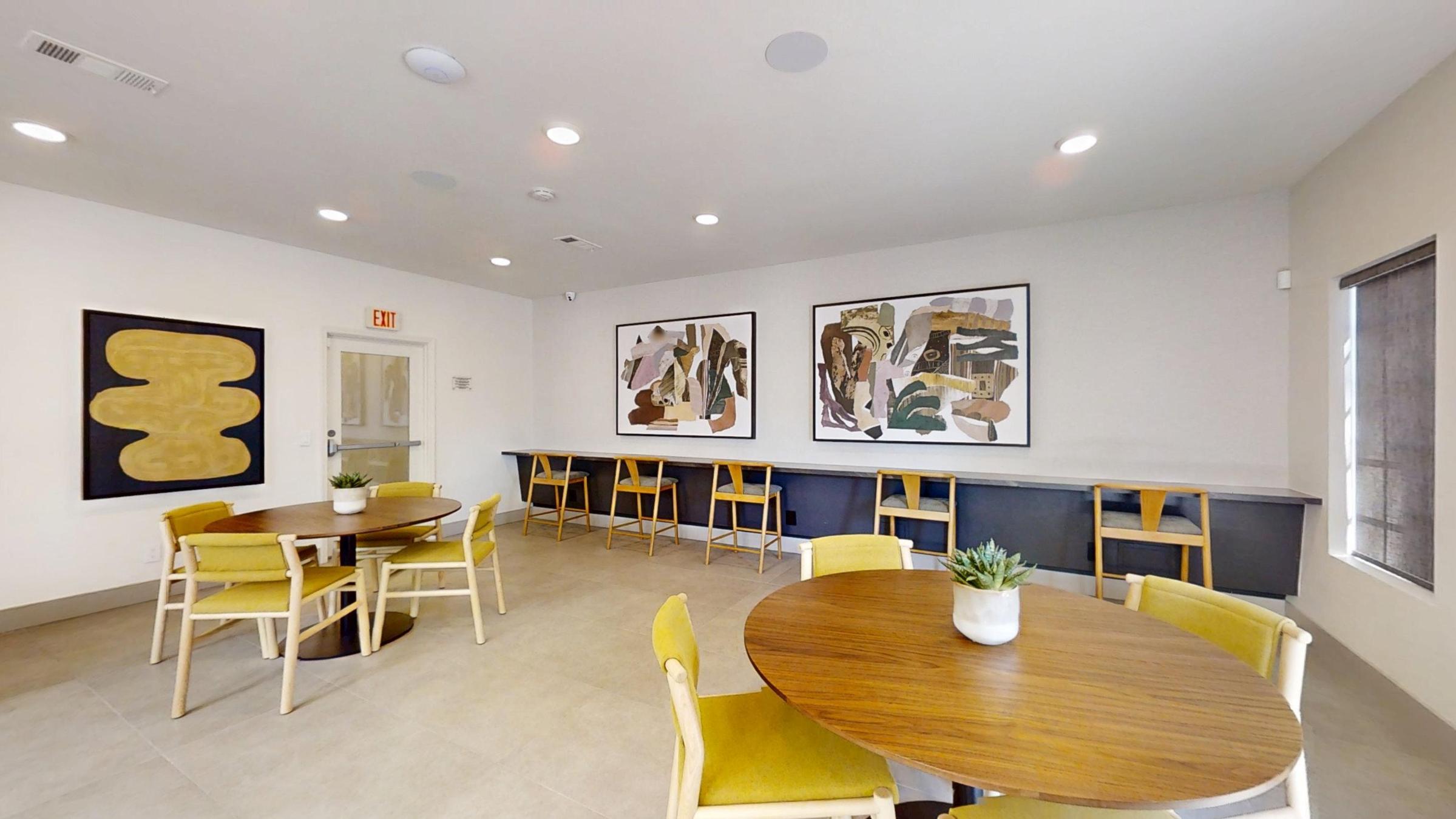 A modern lounge area features two round wooden tables surrounded by light-colored chairs. A long counter with high stools runs along one wall, and two abstract artworks hang above. The space has neutral-toned walls and ample lighting, creating a welcoming atmosphere. A small potted plant is on one table.
