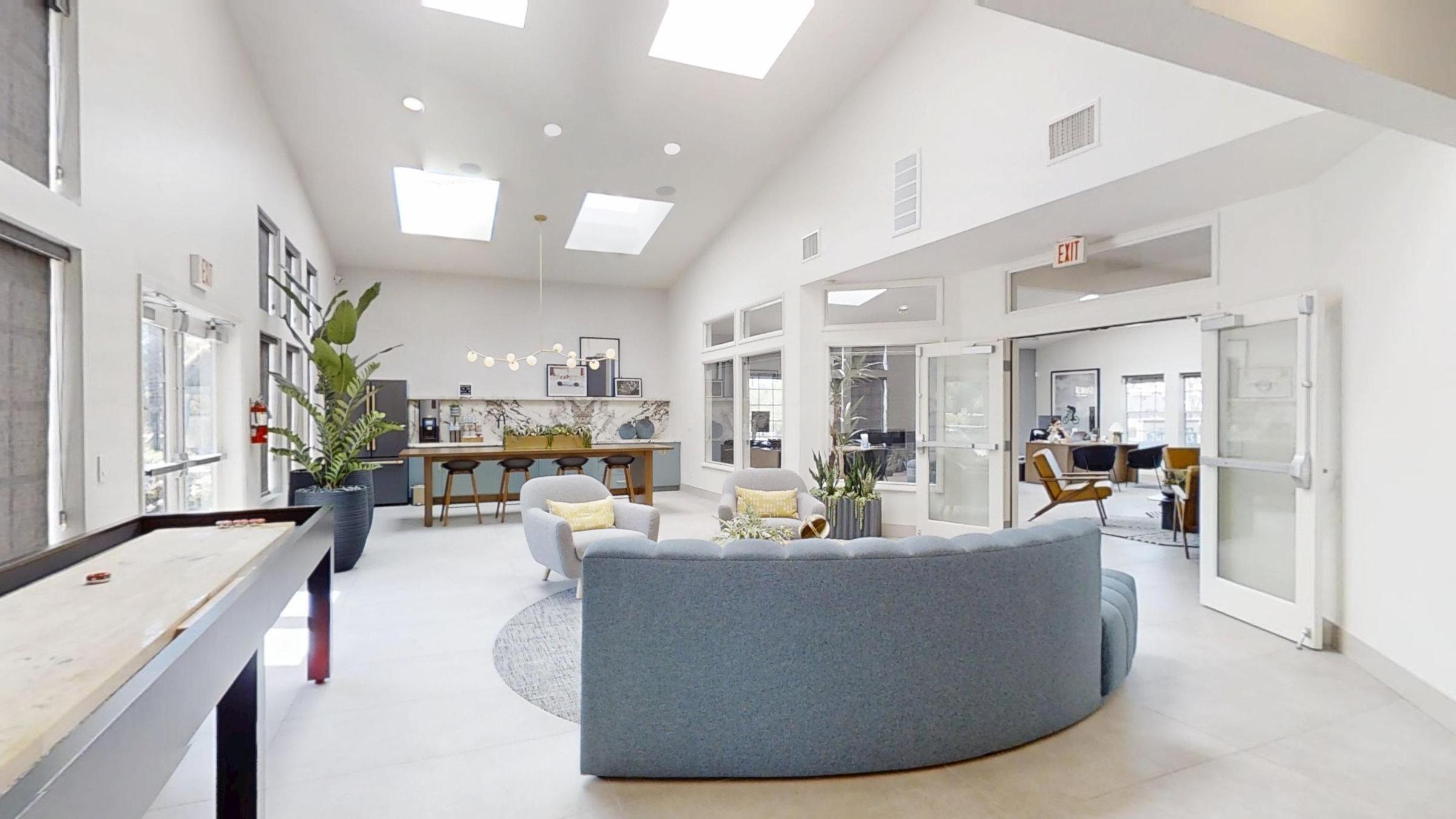 A spacious and modern lounge area with large skylights, featuring a curved blue couch, potted plants, and a coffee table. In the background, there are tables and chairs, as well as doorways leading to other rooms. The overall decor is bright and inviting, with neutral tones and contemporary furnishings.