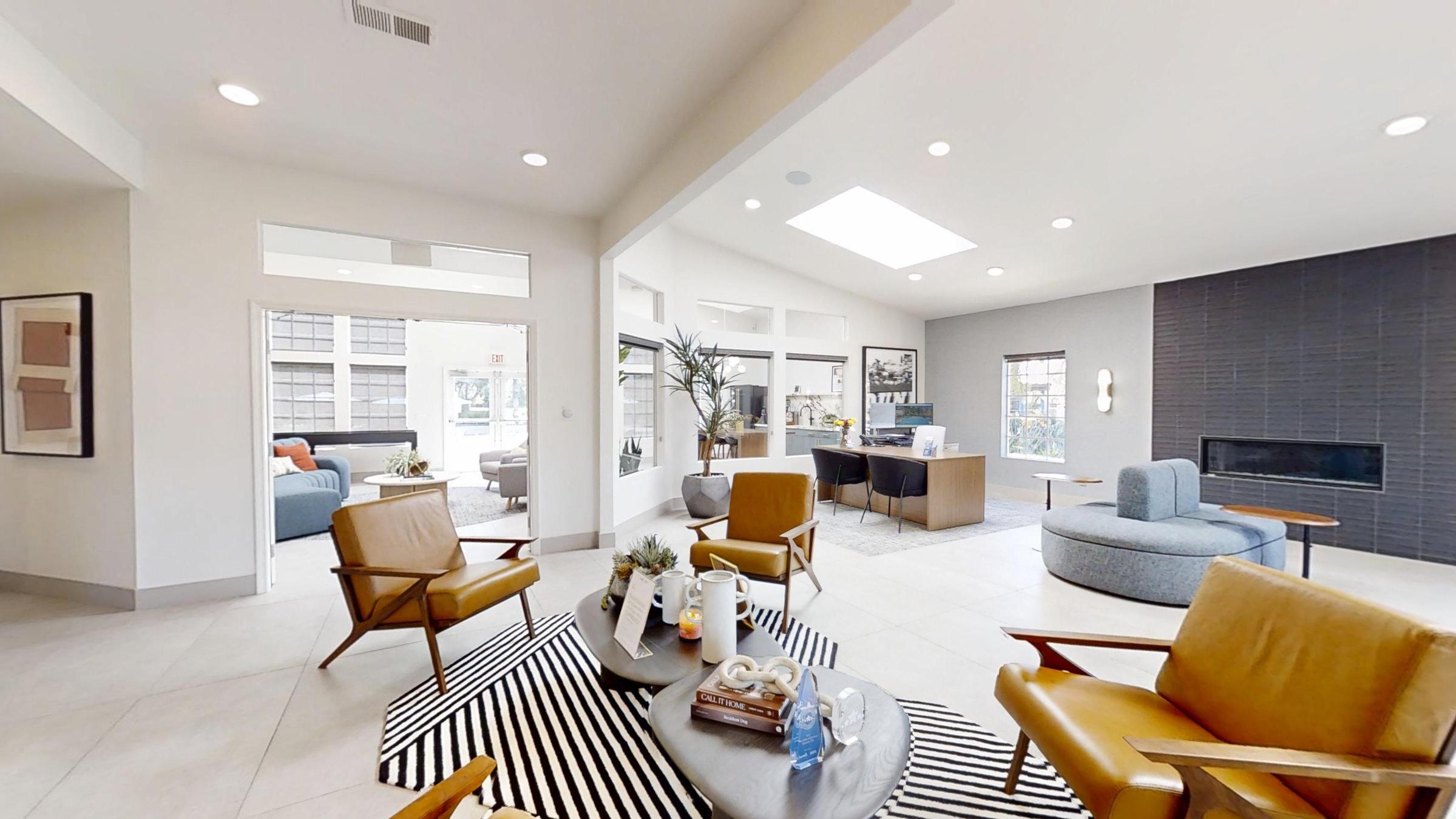A modern living area featuring two brown armchairs, a striped rug, a coffee table with decorative items, and a cozy atmosphere. Large windows provide natural light, and there are plants for decoration. In the background, a casual seating area with a blue sofa and a hearth can be seen.