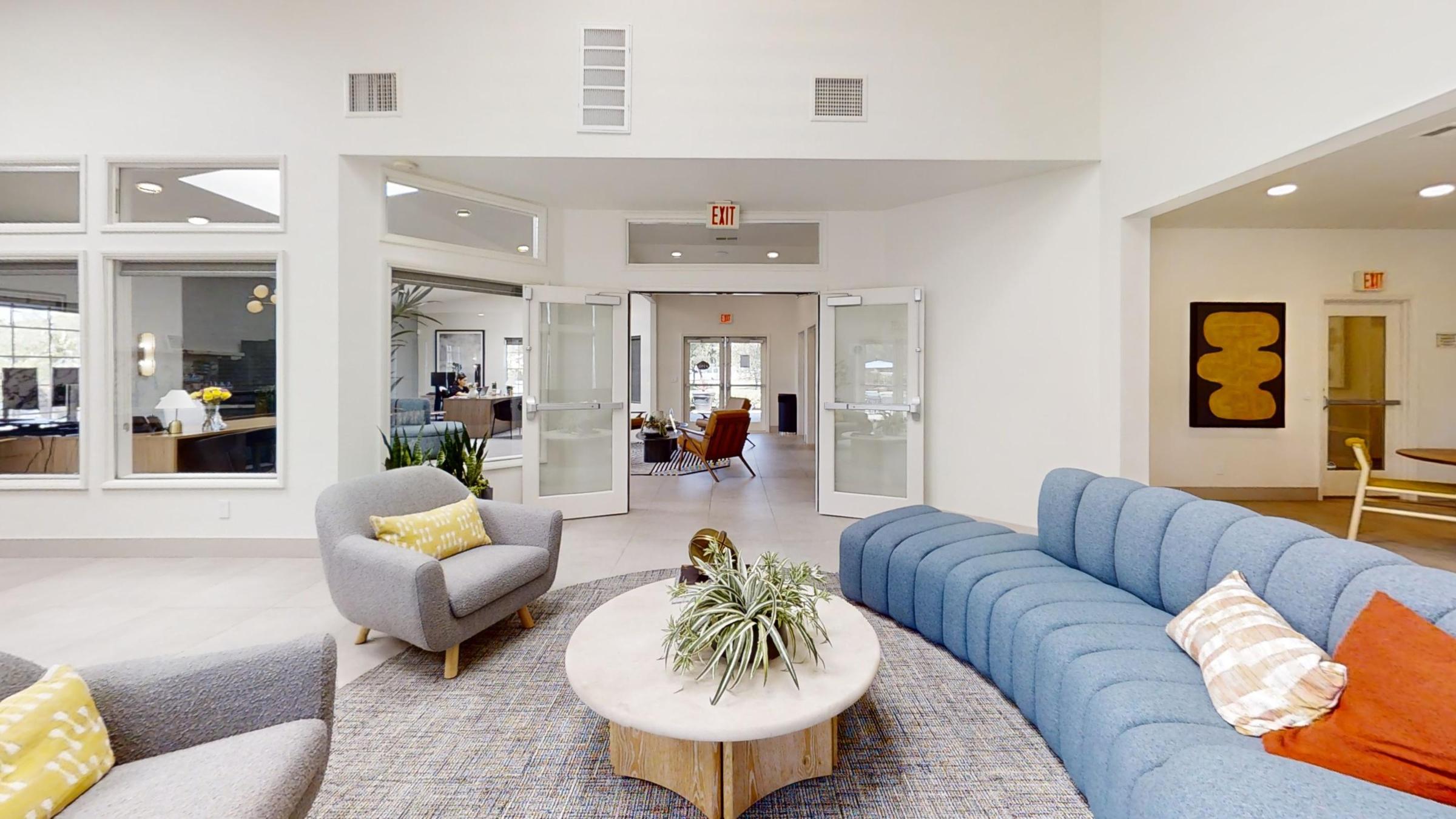 A modern, inviting common area featuring a blue sectional sofa, a round coffee table with a plant centerpiece, and a textured area rug. Large windows allow natural light, while doors lead to a well-lit dining area with stylish furnishings in the background.