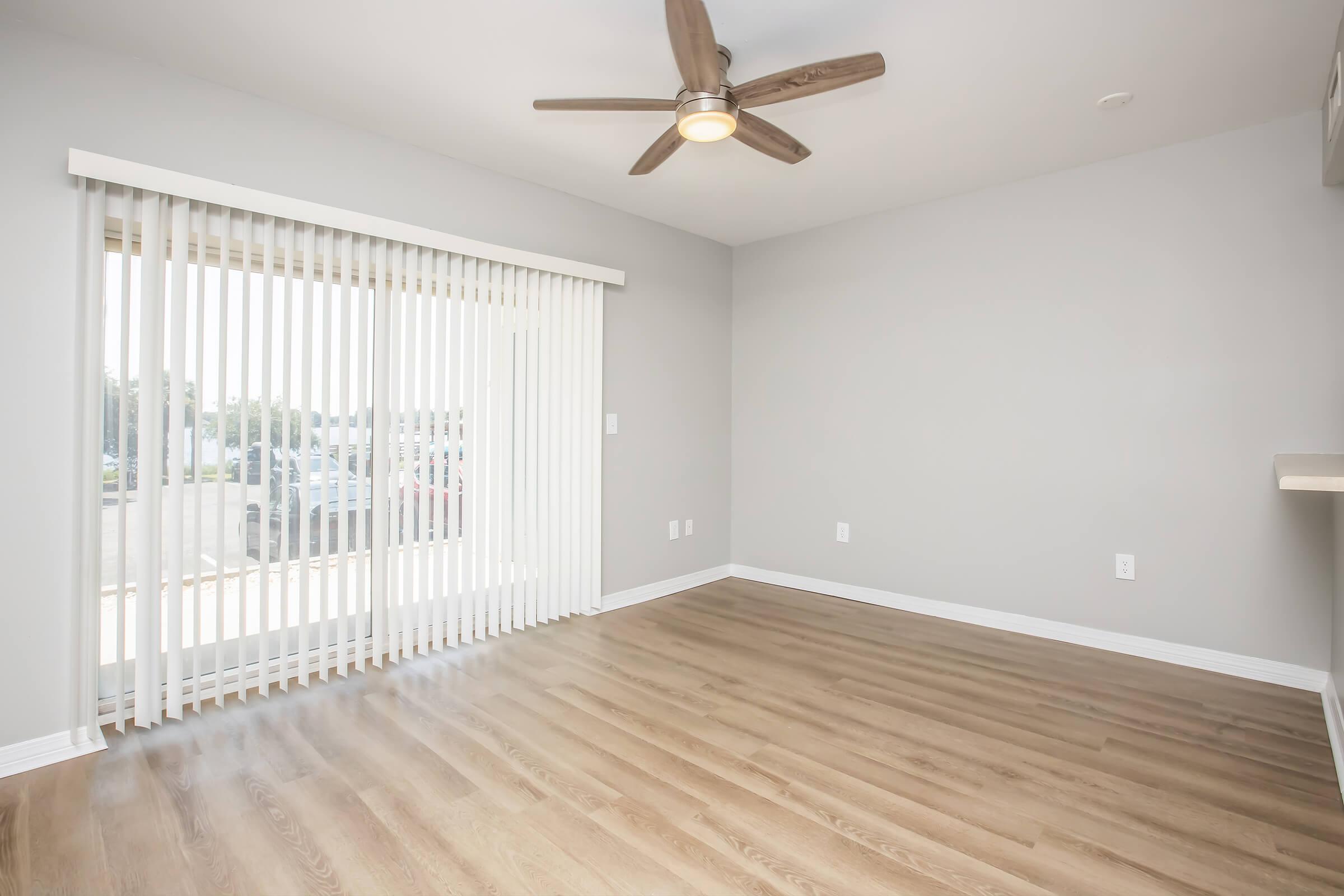 A spacious, empty room with light gray walls and wooden flooring. A ceiling fan is visible above, and large sliding glass doors with vertical blinds allow natural light to enter. The room is unfurnished, creating a clean, open atmosphere.