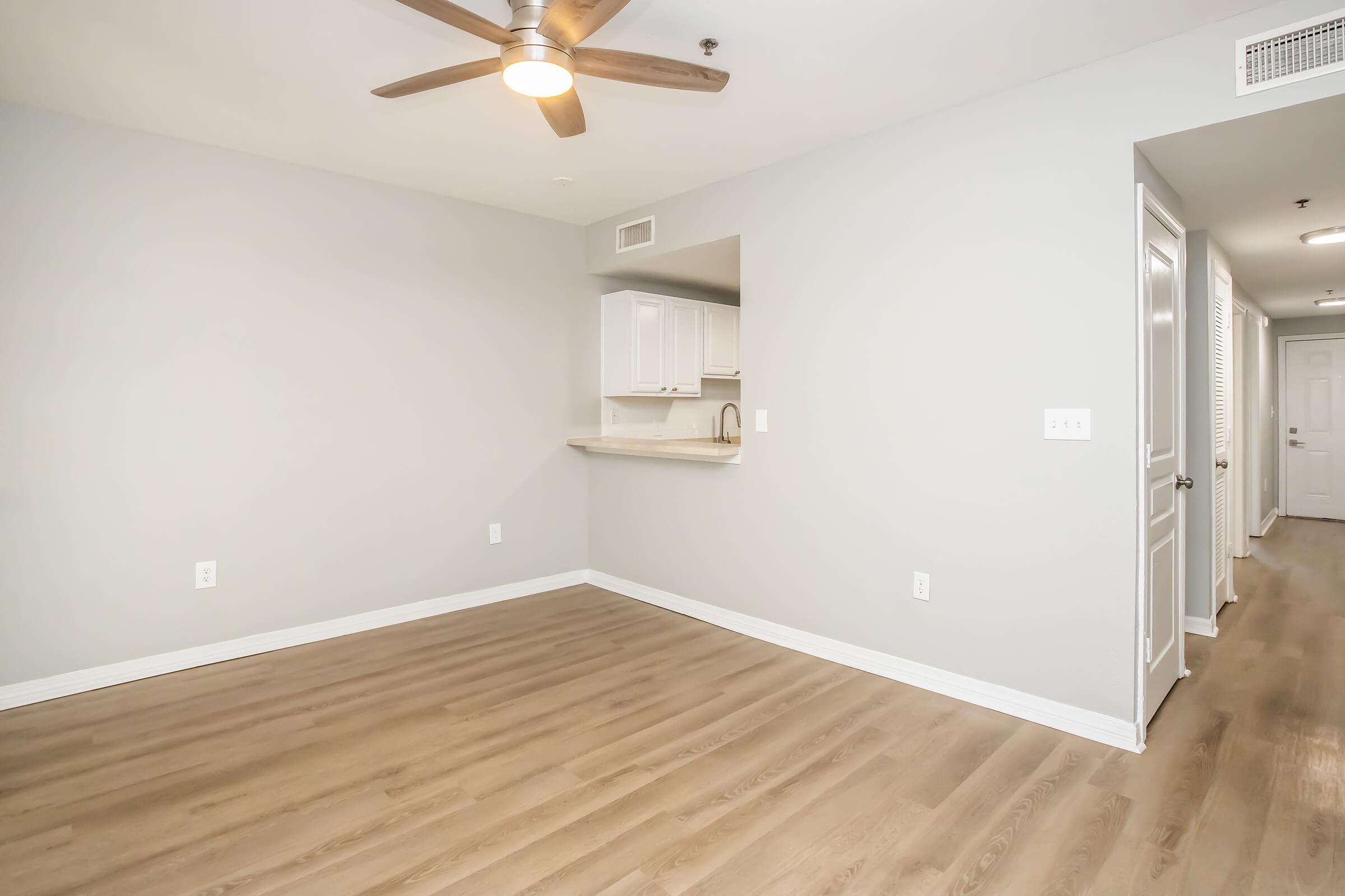 A spacious, empty room with light gray walls and new hardwood flooring. There is a ceiling fan overhead and a small open kitchen area with white cabinetry visible on the left. The right side features a hallway leading to additional rooms, enhancing the room's bright and airy feel.