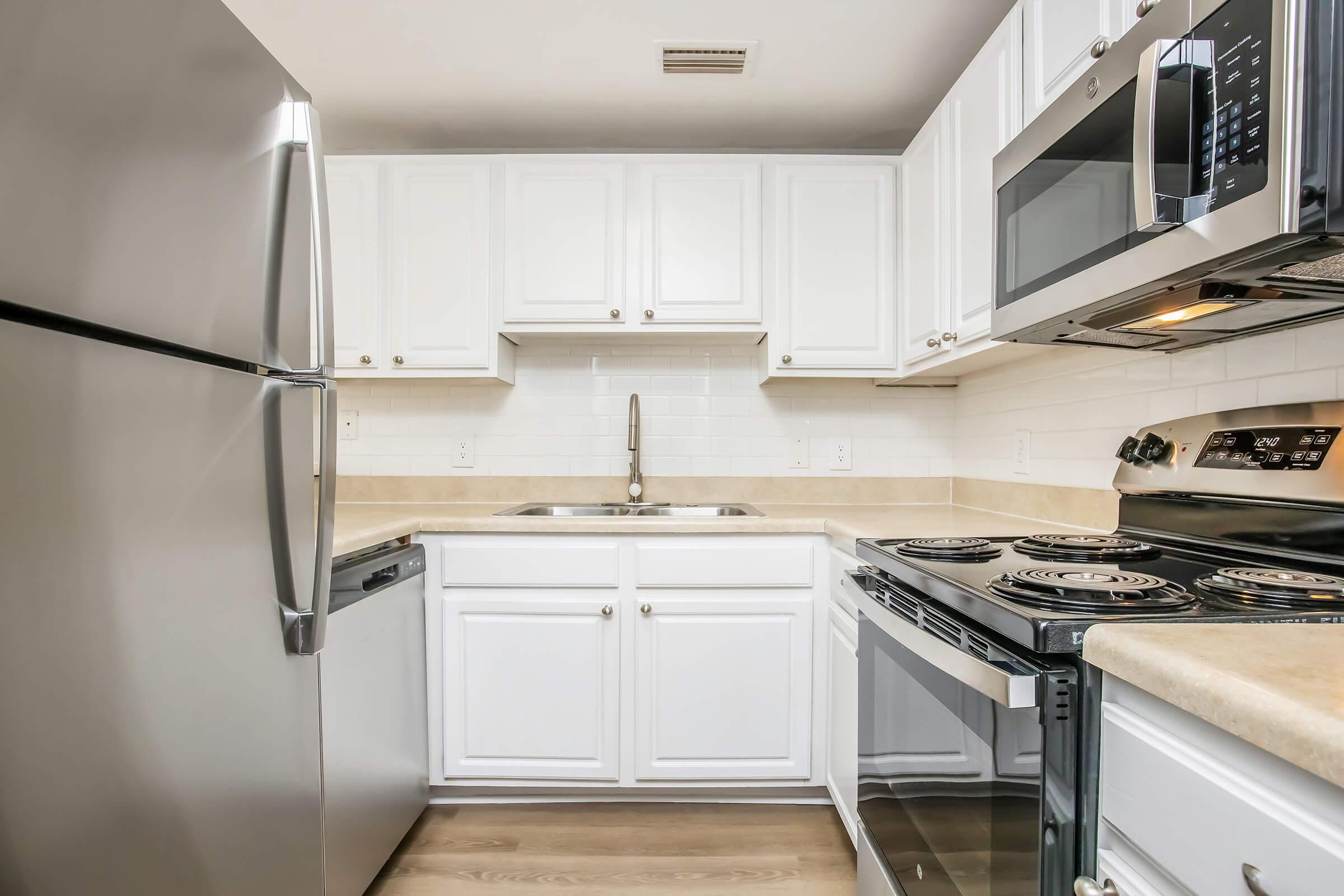 A modern kitchen featuring white cabinetry, stainless steel appliances including a refrigerator, dishwasher, and microwave. The countertop is a light beige color, and the space has a clean, organized layout with a sink and a four-burner stove. The flooring is light wood.