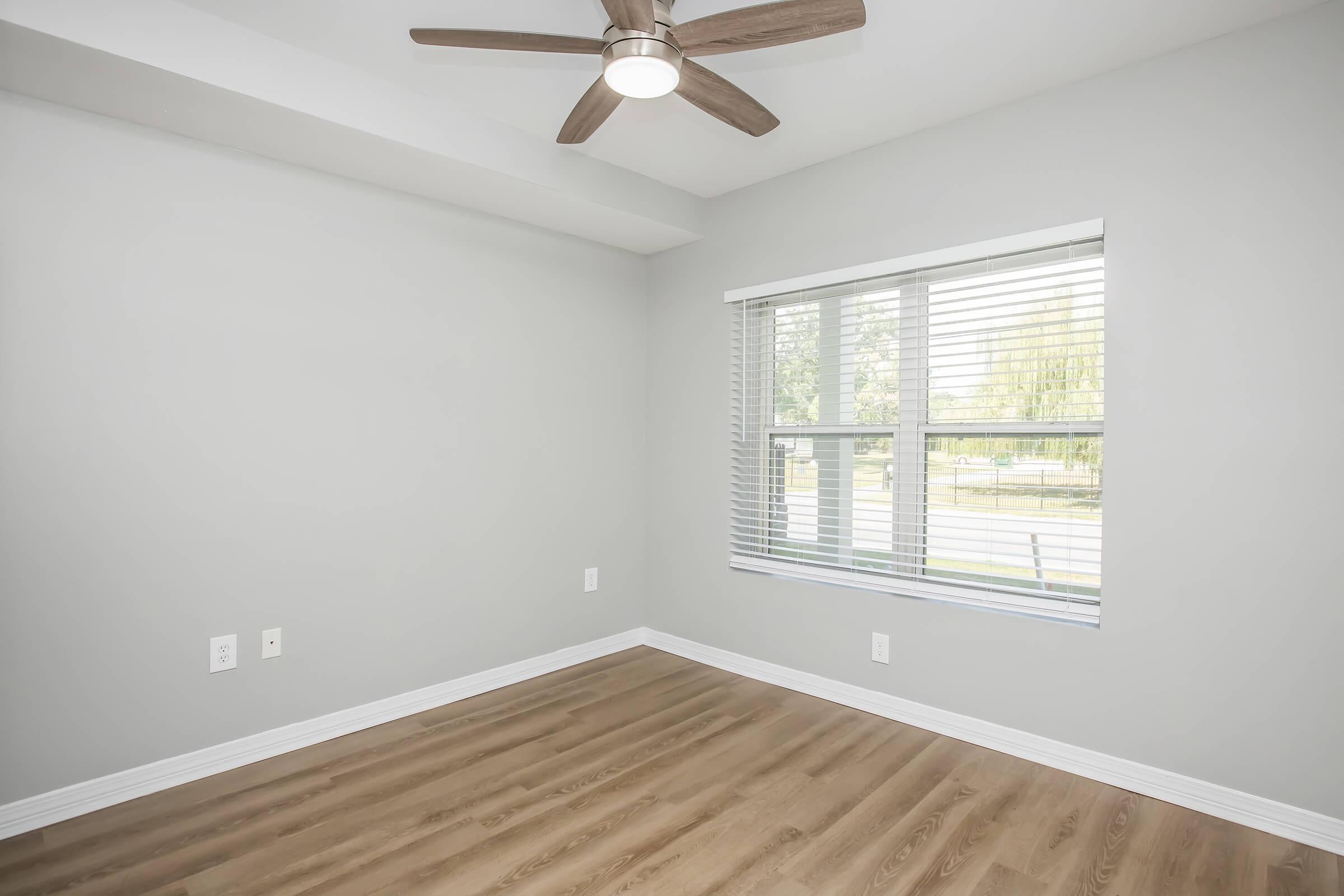 A spacious empty room featuring light grey walls, a ceiling fan, and a large window with blinds. The floor is made of light wood, and there's natural light coming in from outside. The room appears clean and ready for furnishings.