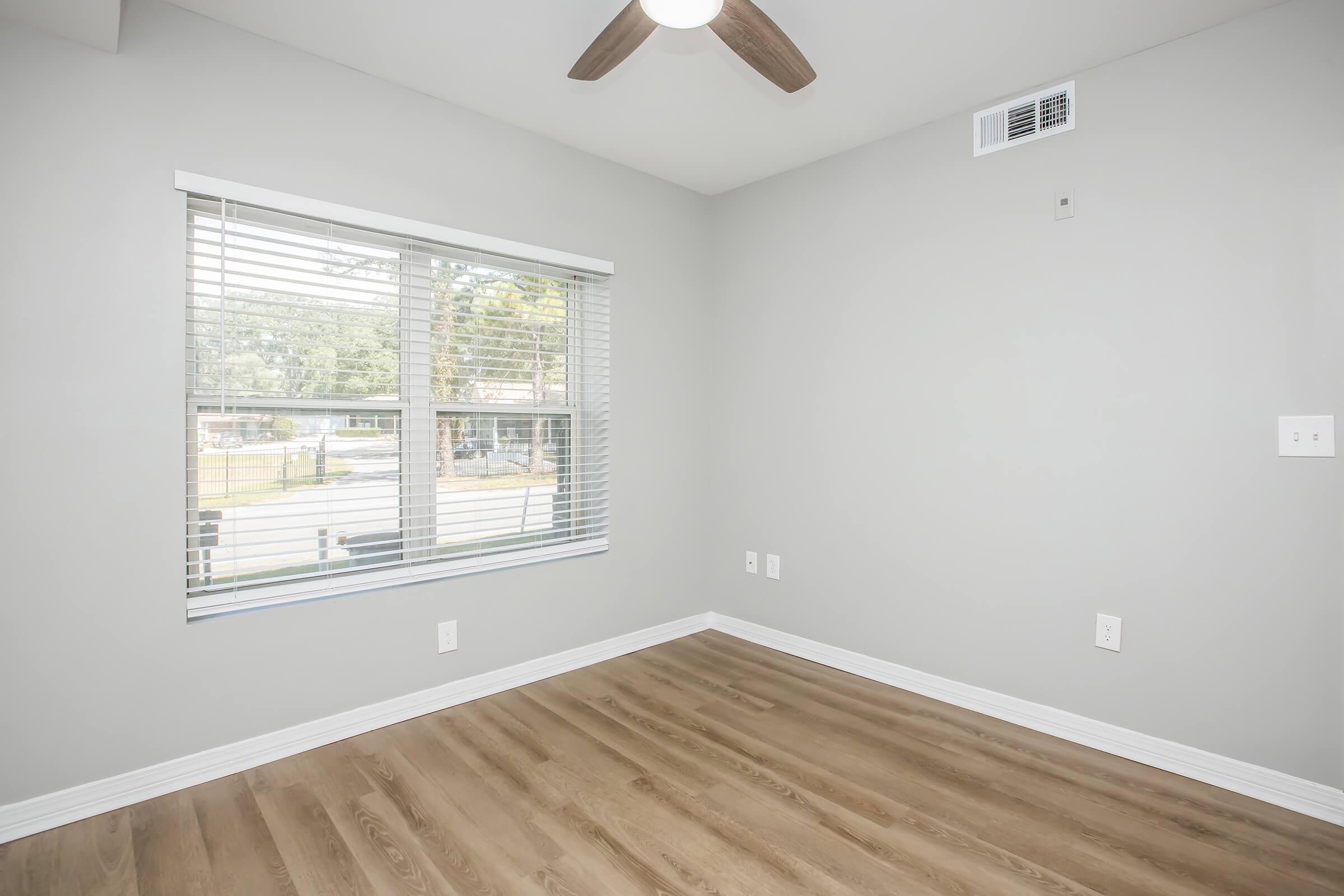 A bright, minimalistic room featuring a ceiling fan and large window with blinds. The walls are painted light gray, and the floor is wooden with a natural finish. There are electrical outlets on the walls, and the overall ambiance is clean and modern.