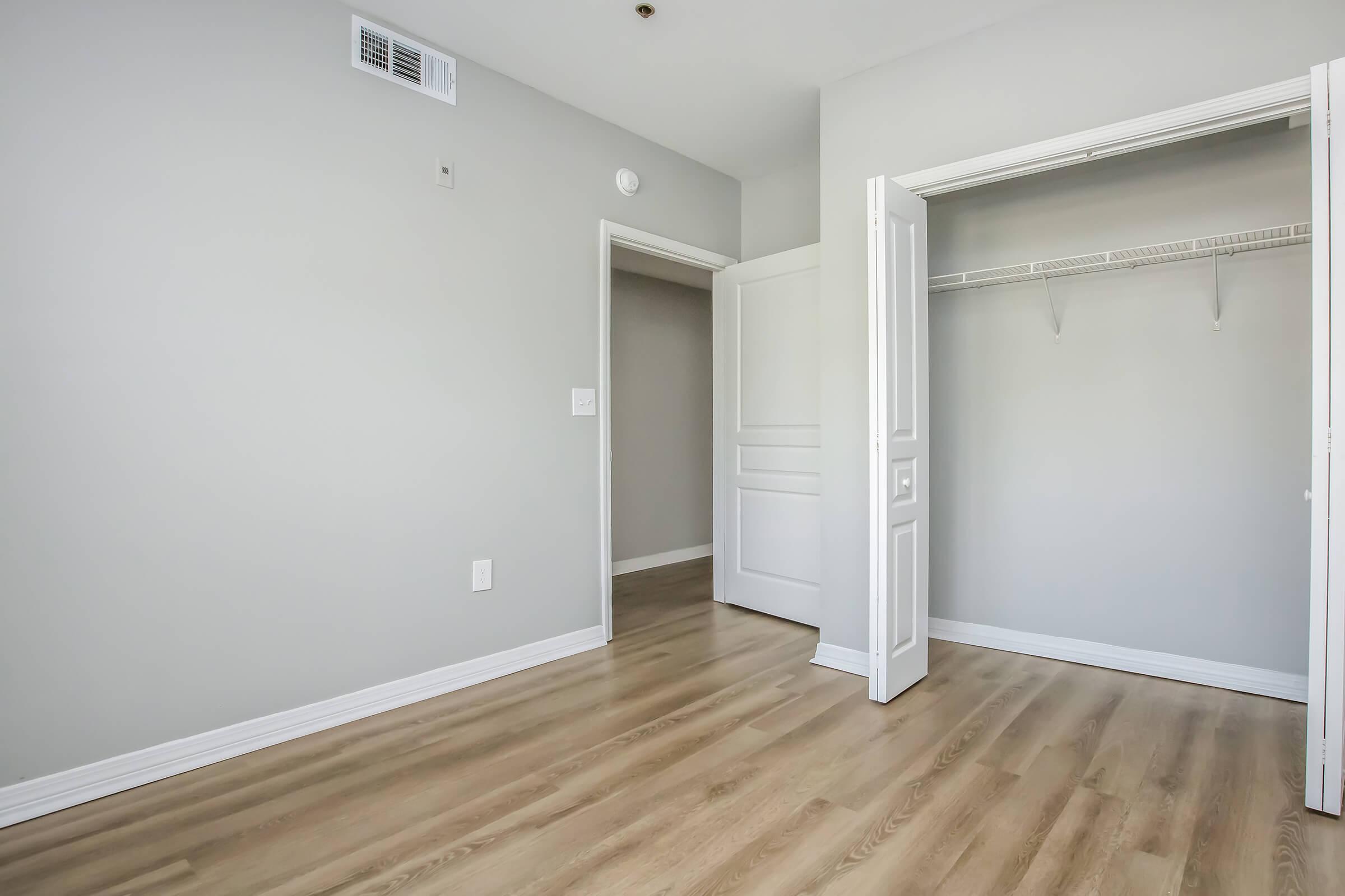 A spacious, empty room with light gray walls and hardwood flooring. One side features an open closet with a shelf, while another side has a closed door leading to a hallway or another room. Natural light may be coming in, enhancing the airy feel of the space.