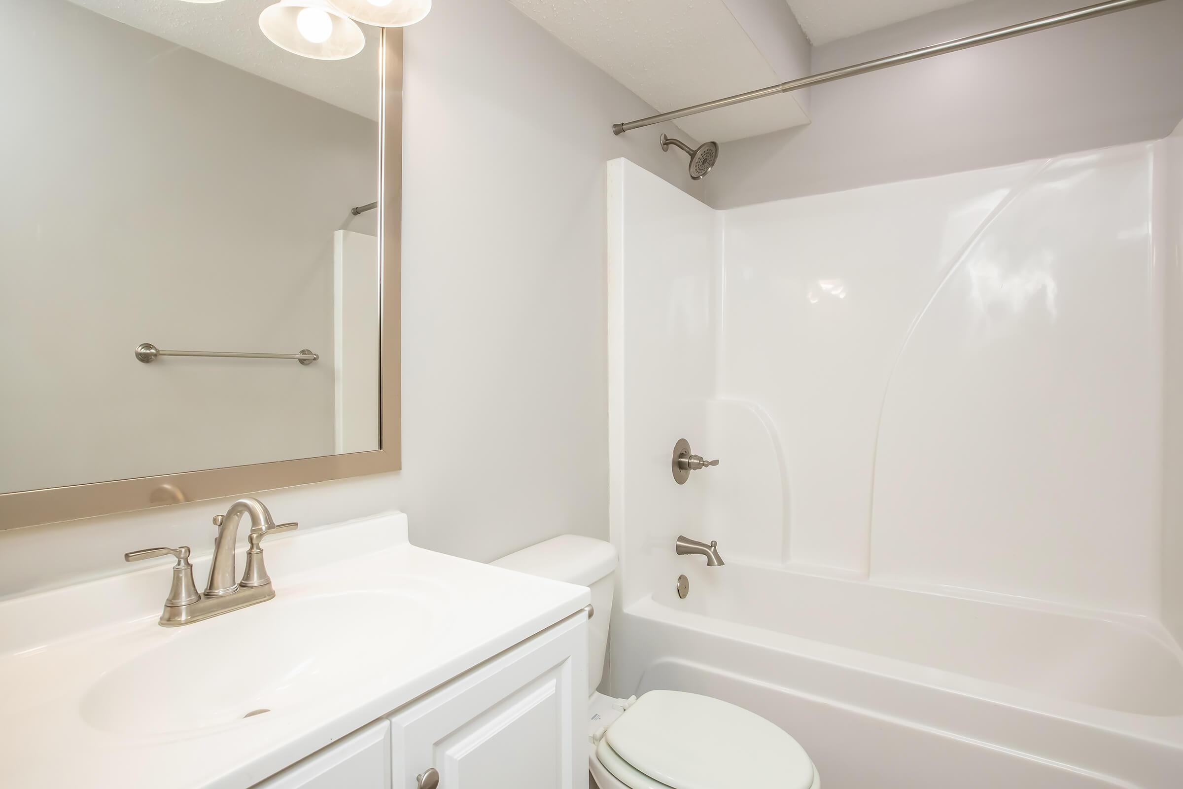 A clean and modern bathroom featuring a white bathtub and shower combination, a white sink with brushed nickel faucets, a large mirror, and neutral-colored walls. The space is well-lit with overhead lighting and has simple, minimalist design elements.