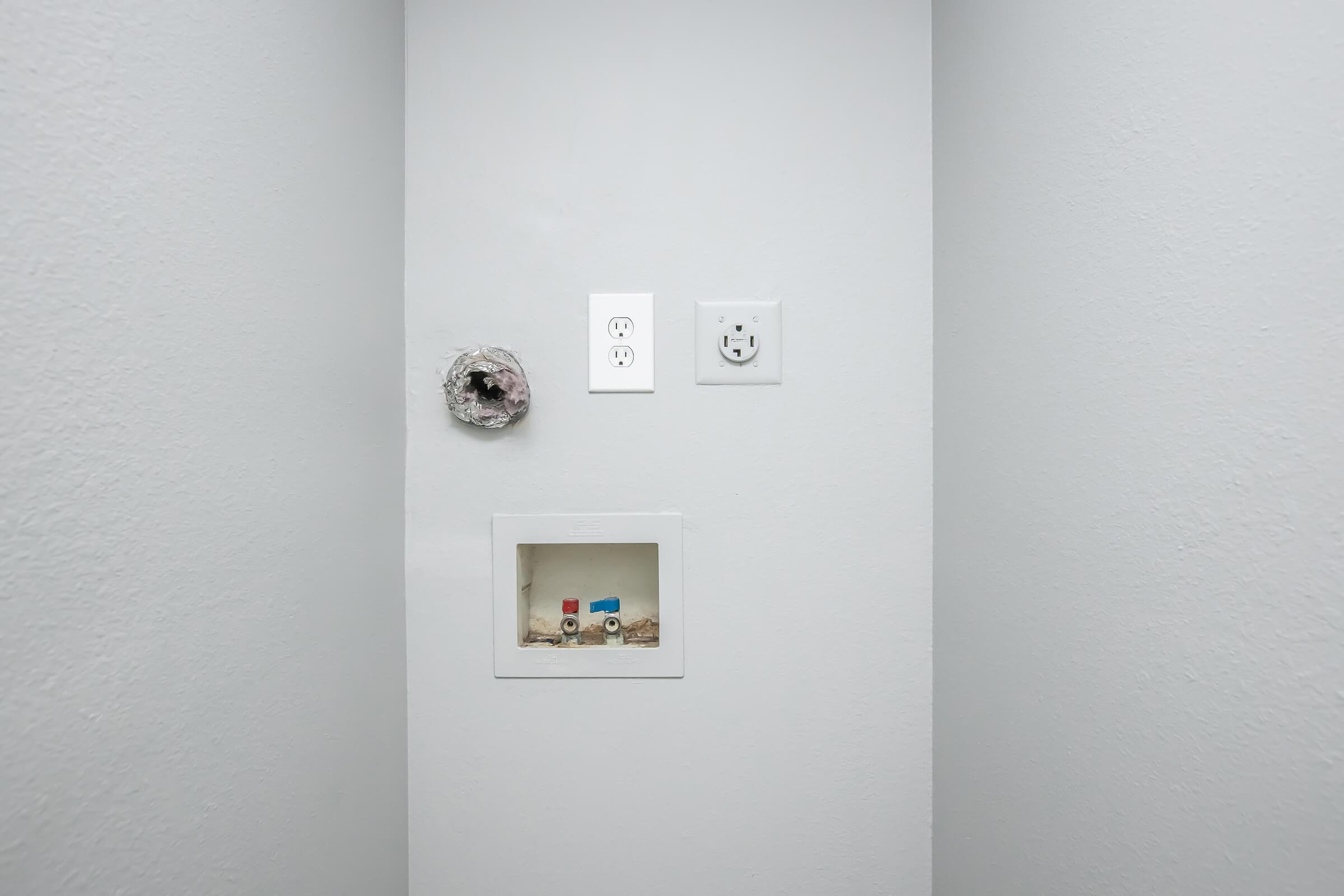 A plain white wall featuring two electrical outlets, one standard and one with a cover plate. Below the outlets, there is a small recessed area with a few colorful objects, possibly tools or toiletries, placed inside a shallow container. The overall appearance is minimalistic and clean.