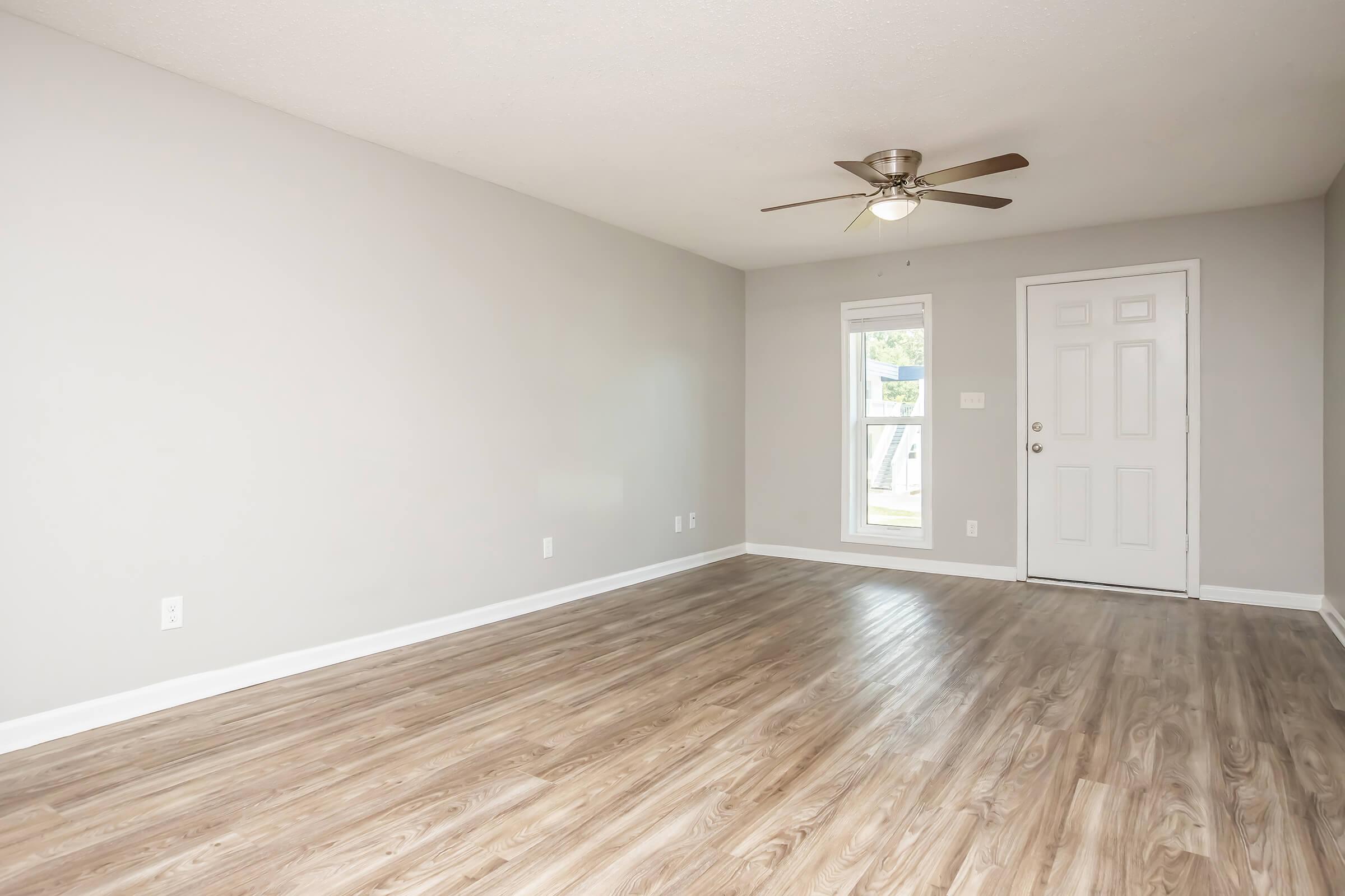 A spacious, empty room with light gray walls and a ceiling fan. It features a large window on one side, allowing natural light, and a front door with a simple design. The floor is covered in light wood laminate, creating a warm and inviting atmosphere.