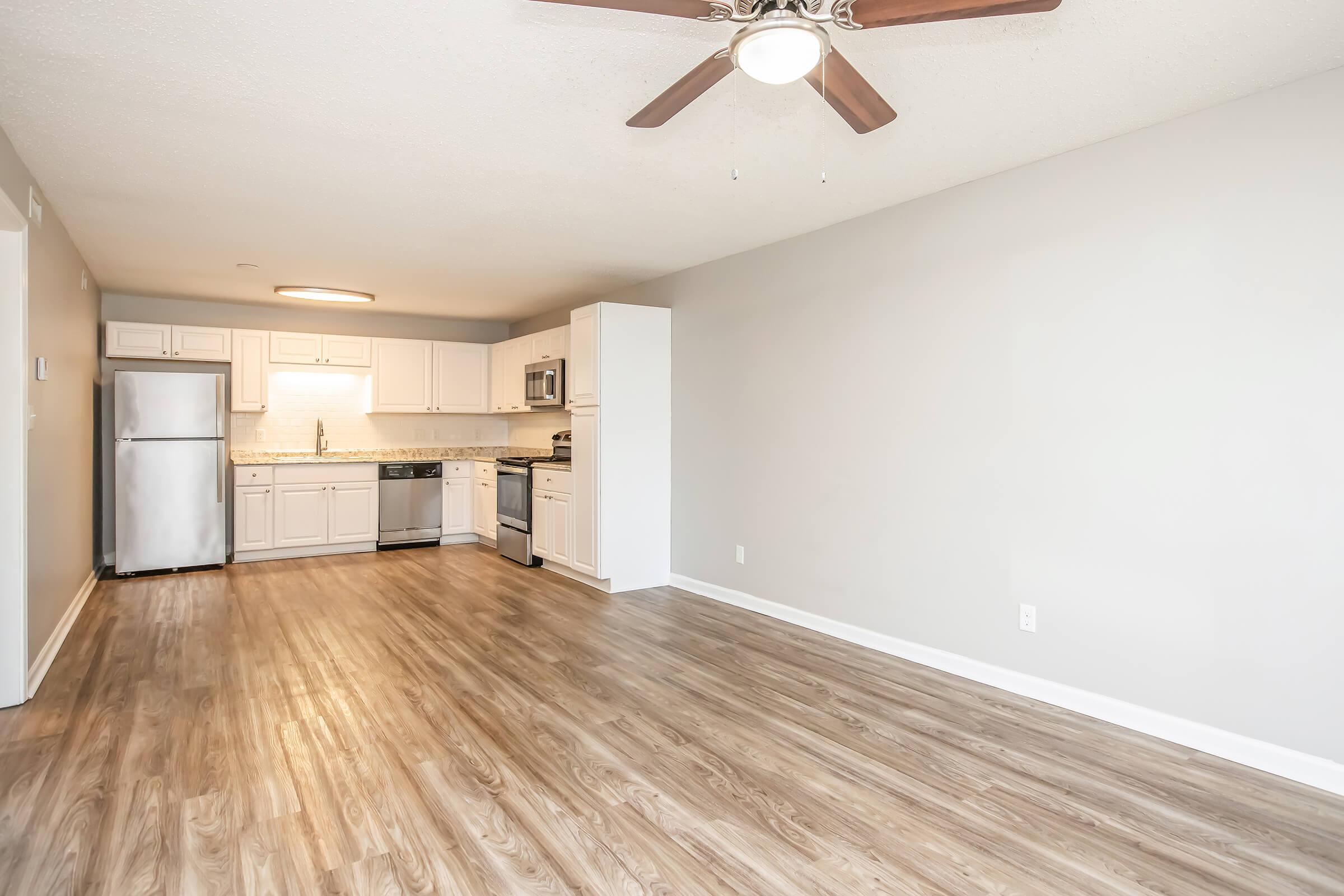 A modern, spacious apartment living area featuring a kitchen with stainless steel appliances, white cabinets, and light countertops. The room has laminate flooring, neutral wall colors, and a ceiling fan. Natural light enters through an open space, enhancing the inviting atmosphere of the interior.