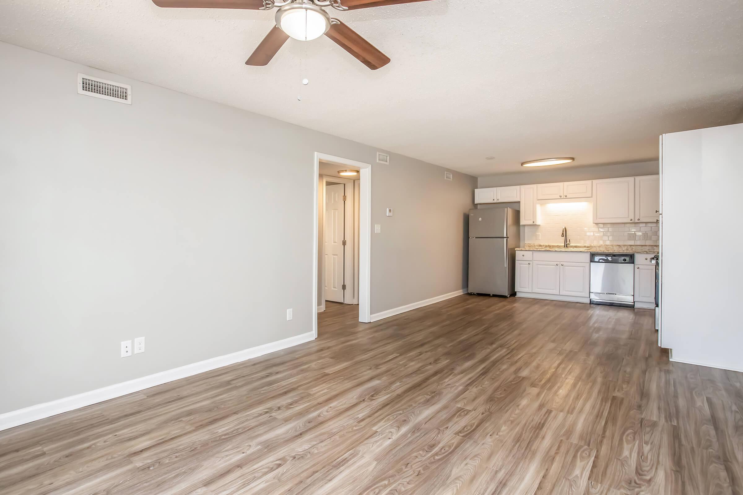 A spacious and modern living area with light-colored walls, a ceiling fan, and laminate flooring. The kitchen features white cabinetry, stainless steel appliances, and a tiled backsplash. An open floor plan allows for easy movement between the living area and the kitchen.