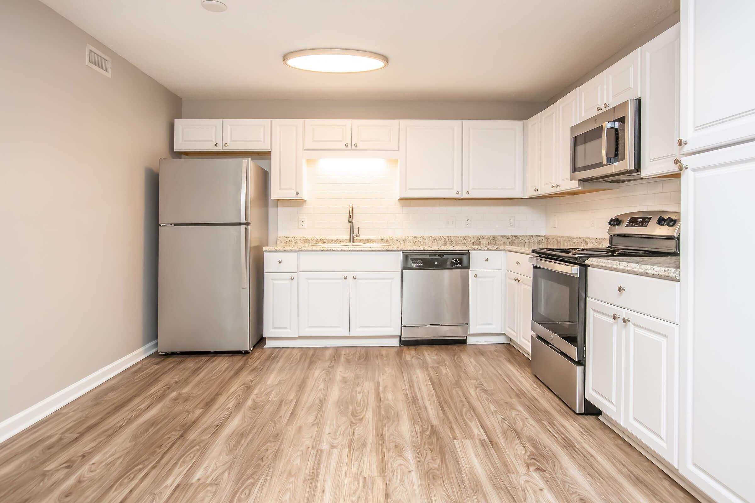 Modern kitchen featuring stainless steel appliances including a refrigerator, dishwasher, and stove. White cabinetry with granite countertops, a single basin sink, and an overhead light fixture. The floor is covered with light wood vinyl, providing a spacious and contemporary feel.