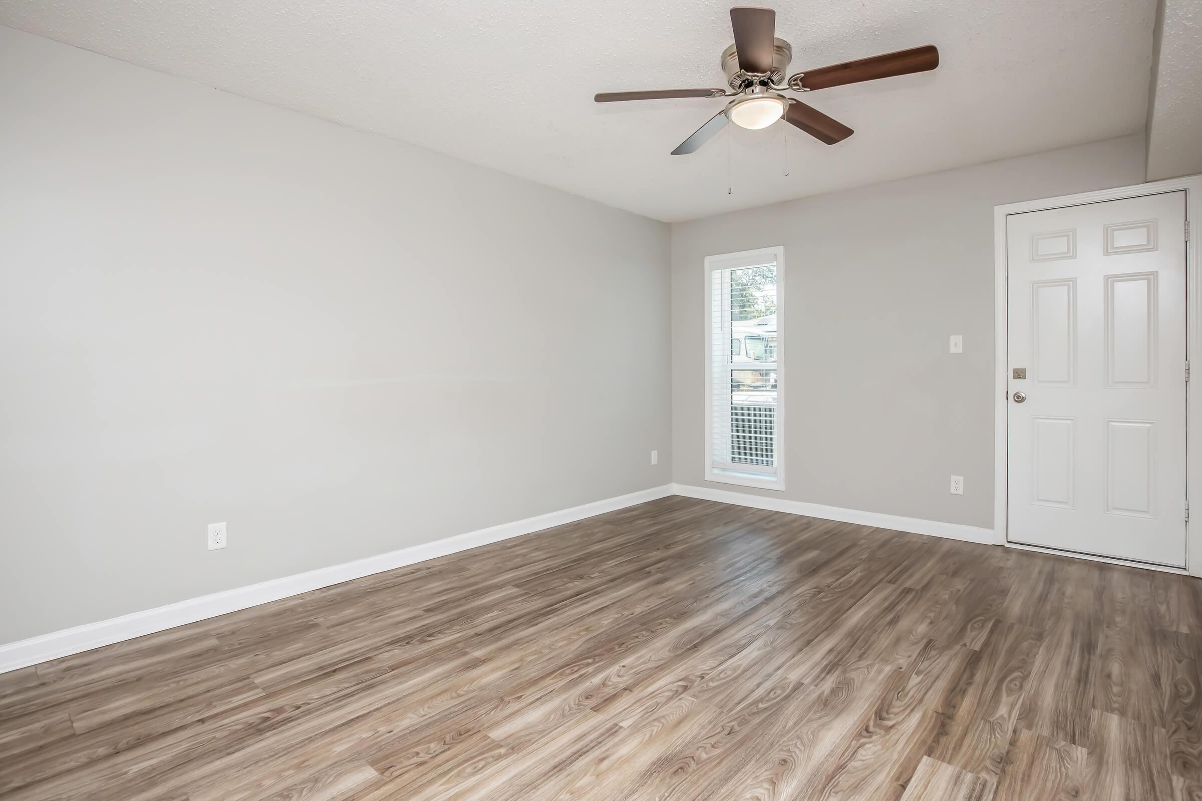 A spacious, well-lit room featuring light gray walls, a ceiling fan, and wood-like flooring. There's a window allowing natural light and a door leading outside. The room is uncluttered, creating a minimalist and inviting atmosphere.
