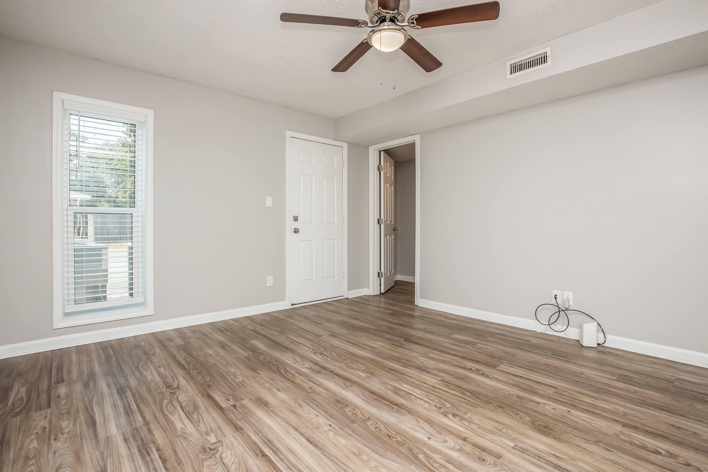 A spacious, empty room with light gray walls and a ceiling fan. The floor has wooden laminate, and there is a window with blinds allowing natural light. A closed door leads to another room, and a white wall outlet is visible on the floor.