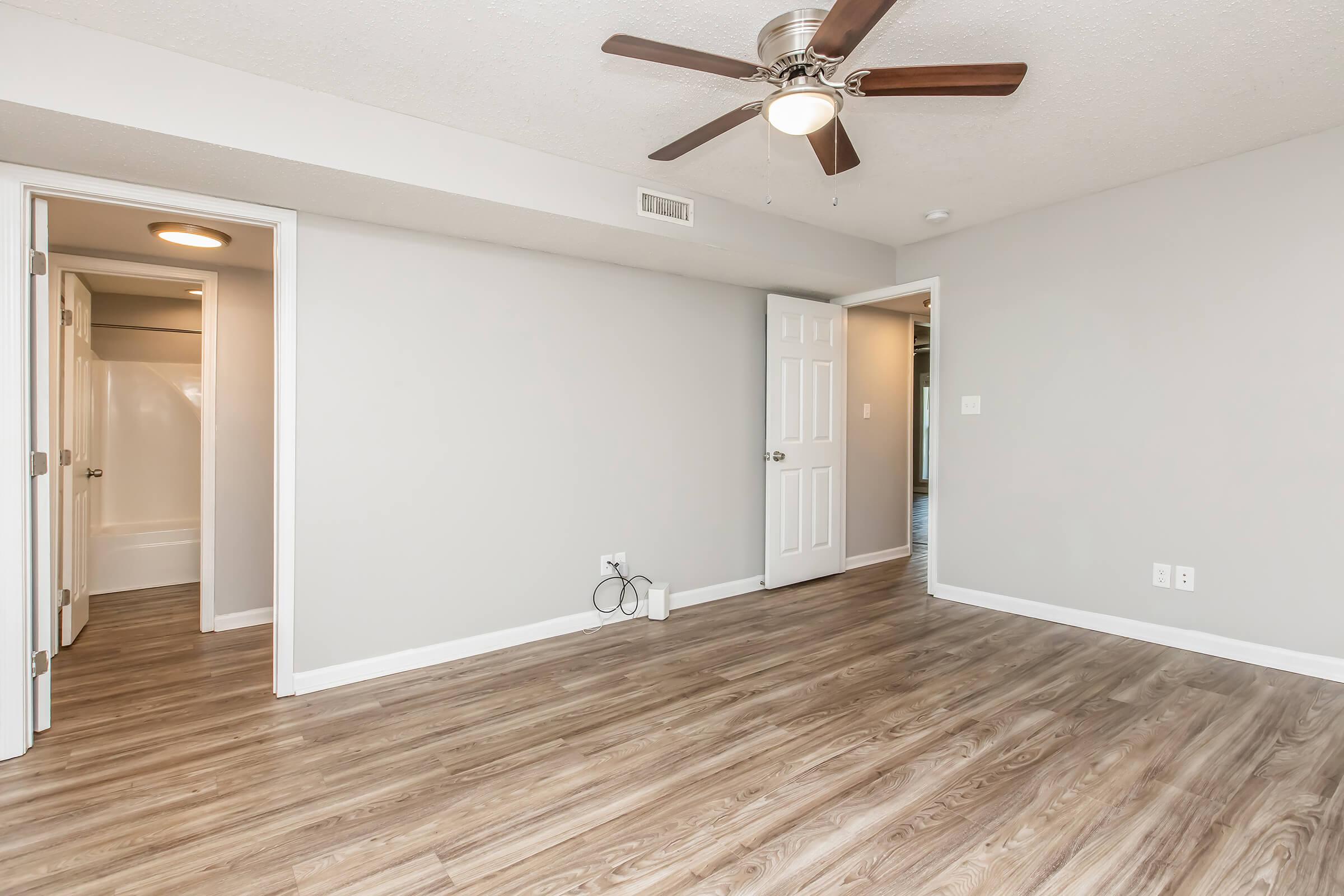 A well-lit, empty room featuring light gray walls and a ceiling fan. The floor is covered with laminate wood, and there are two doorways leading to adjoining spaces. One doorway leads to a bathroom, while the other opens to another room. The space appears modern and ready for furnishing.