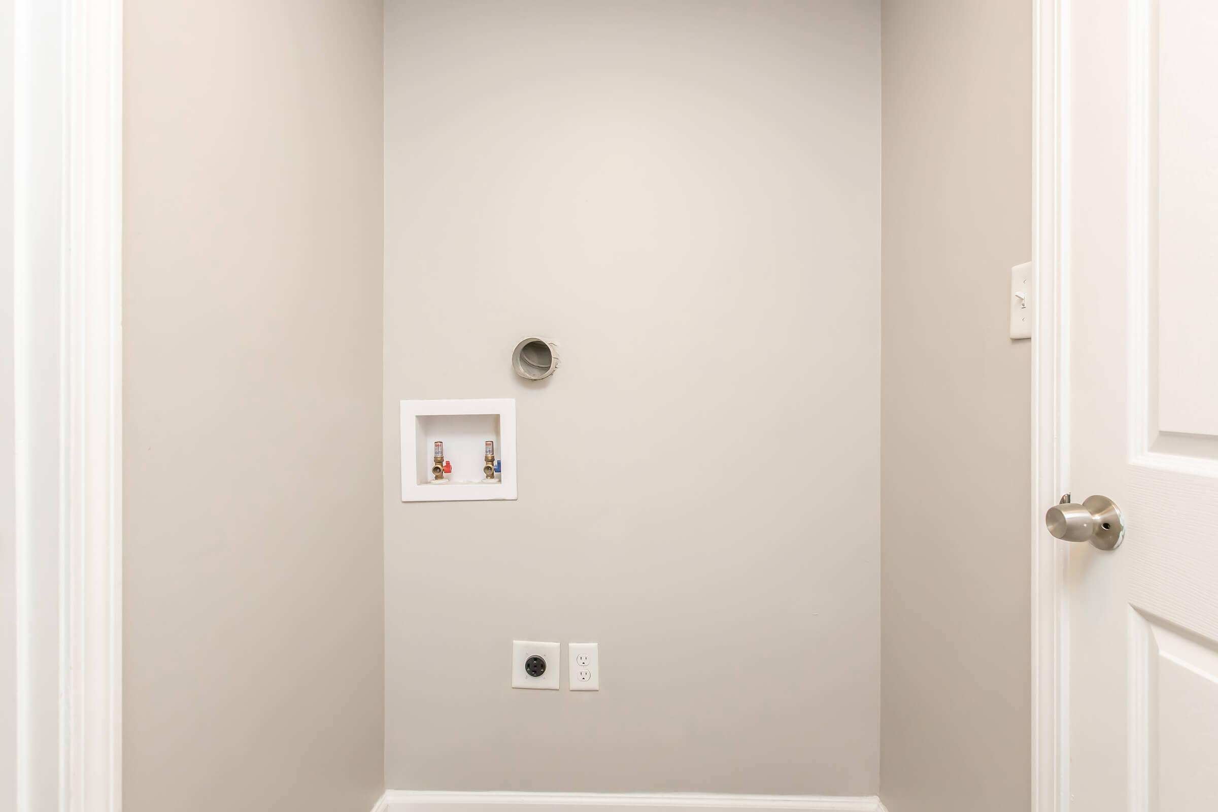 A small, empty room with light gray walls. There is a circular vent or pipe on the wall, and a recessed panel with two small items. A white electrical outlet is visible near the floor, and a door handle is partially shown on the right side. The space appears clean and minimally decorated.