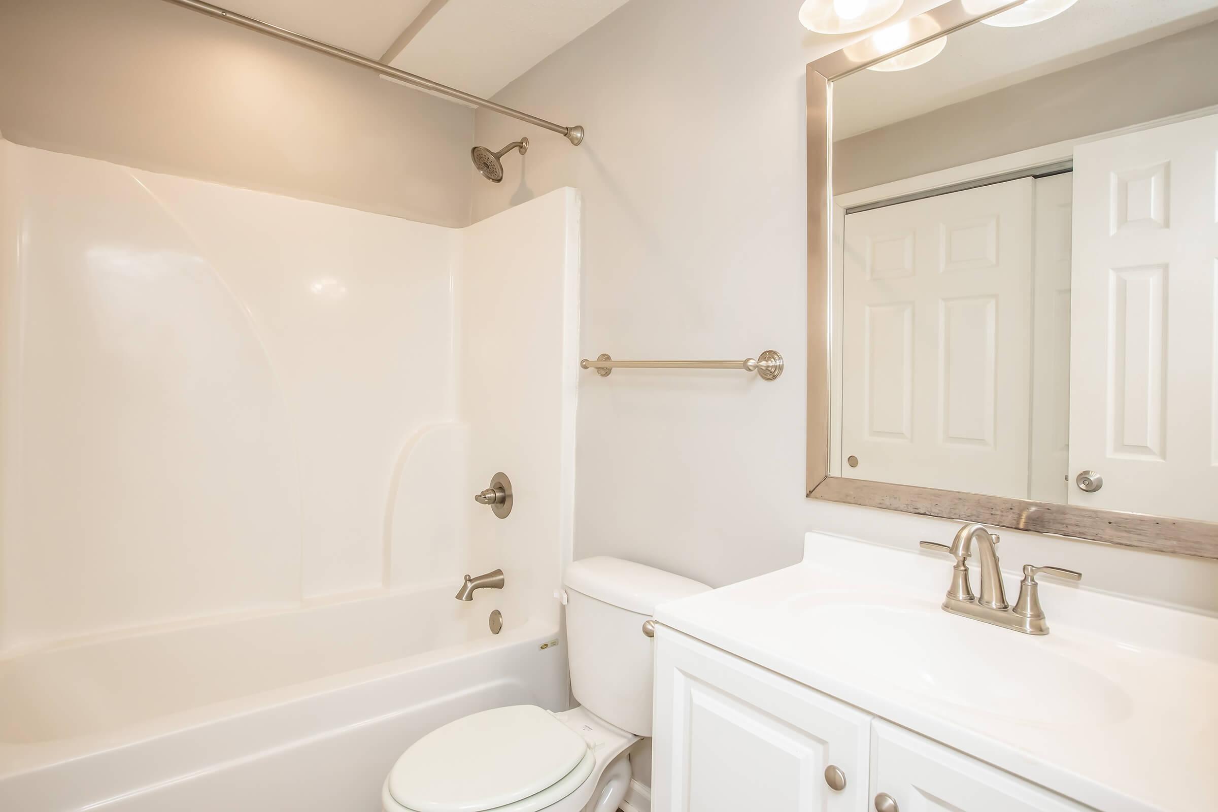 A clean and modern bathroom featuring a white bathtub with a shower, a toilet, and a sleek vanity with a mirror. The walls are painted light gray, and there is a towel bar attached to the wall near the shower. The space appears bright and well-maintained.