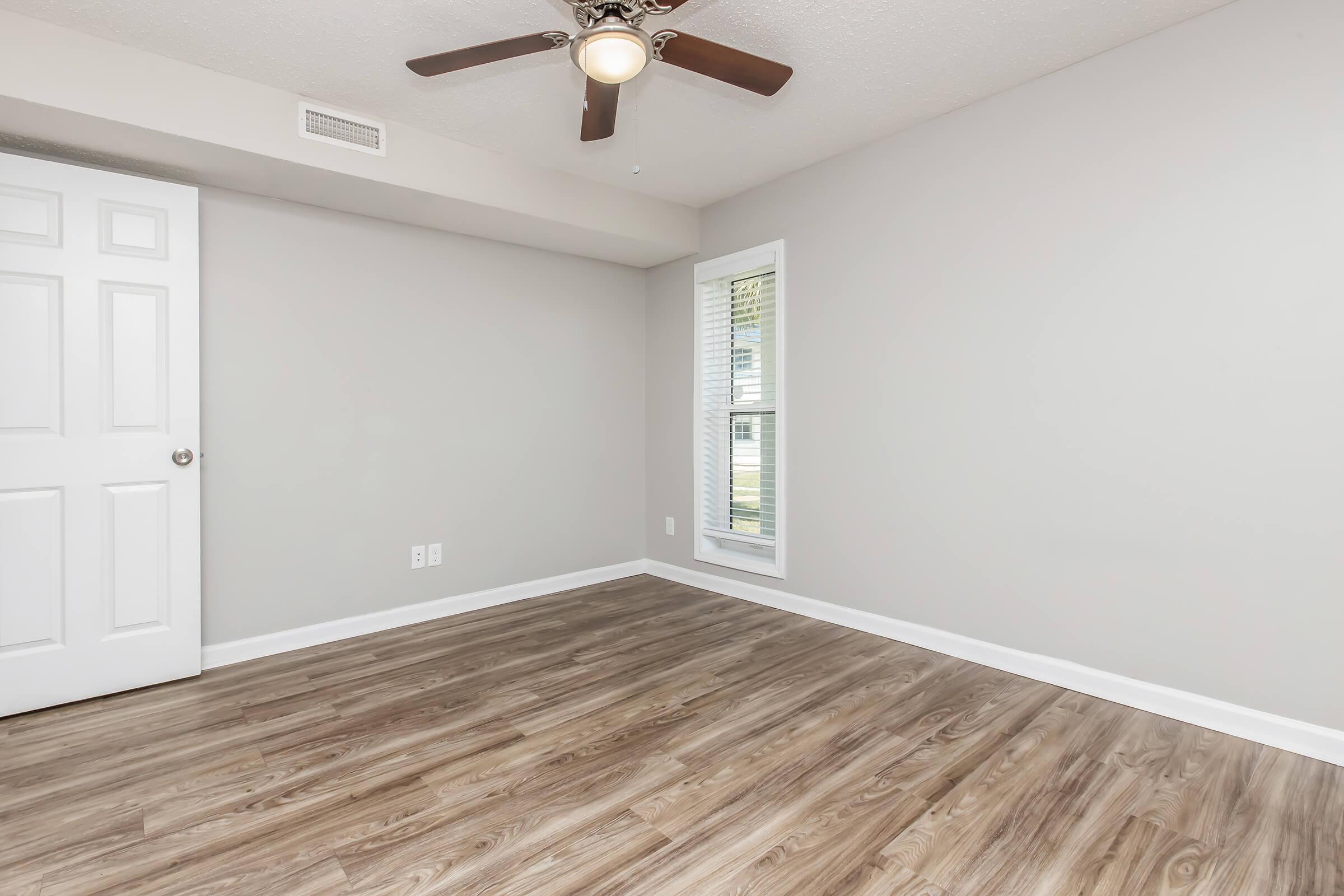 Empty room with light gray walls and a ceiling fan. The floor is wooden laminate, and there is a closet door on the left. A window with a view of the outside is located on the right, allowing natural light to enter the space. The overall atmosphere is bright and inviting.