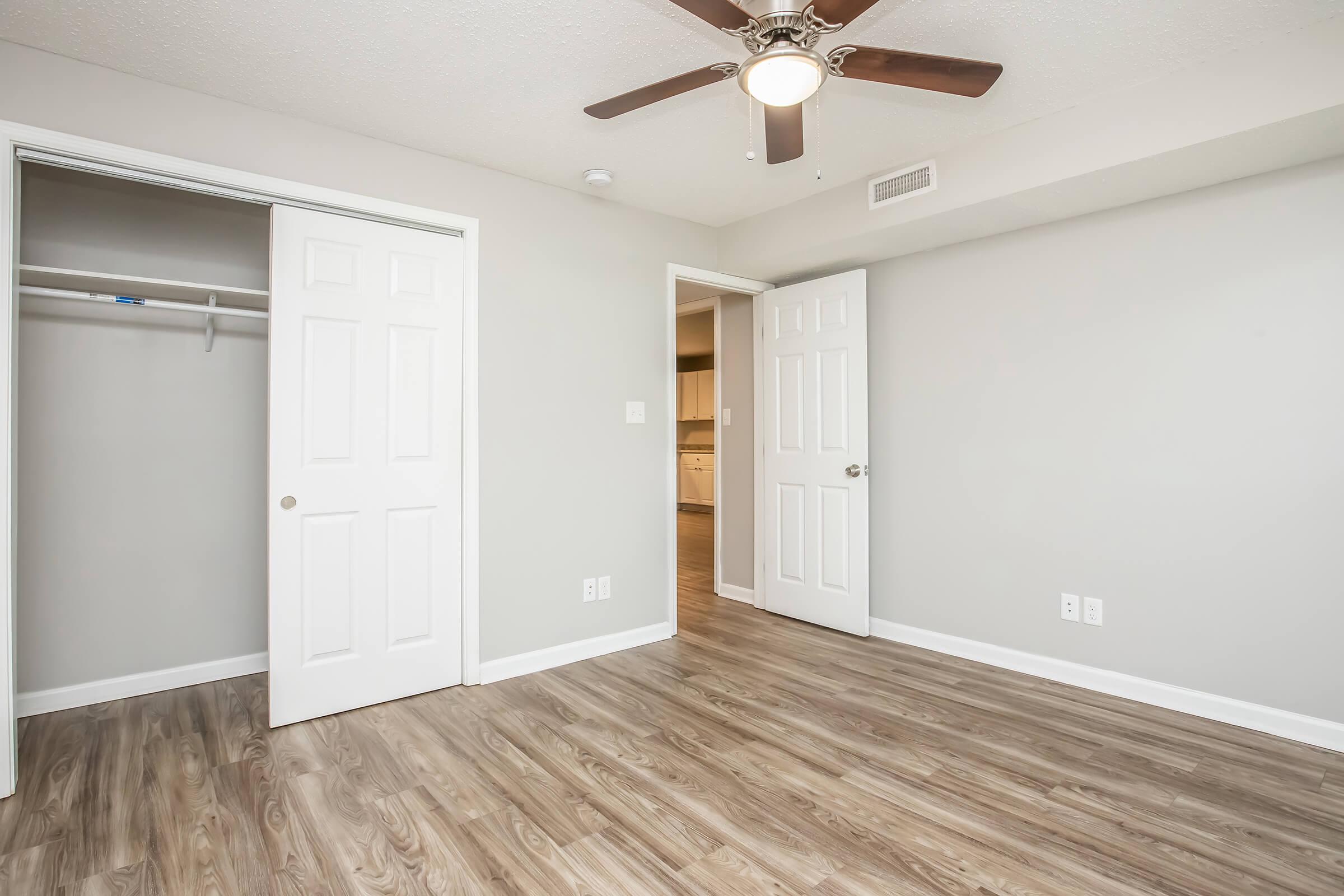 Empty room with light gray walls and wooden flooring. A ceiling fan is mounted on the ceiling, and two white doors are visible: one leading to a closet and the other to another room. The doorway on the right suggests an entrance to a kitchen or living area. Natural light enhances the space.