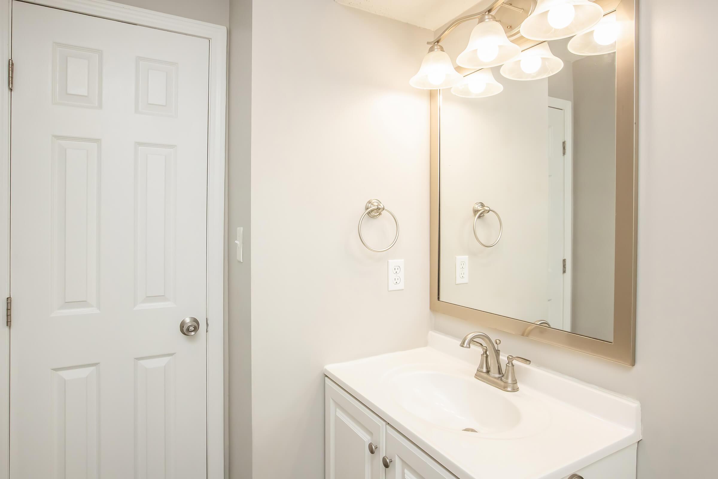 A well-lit bathroom featuring a white vanity with a sink and a mirror above it. There is a stylish faucet and a towel ring on the wall. A closed white door is visible, and the walls are painted in a light color, creating a clean and modern look.