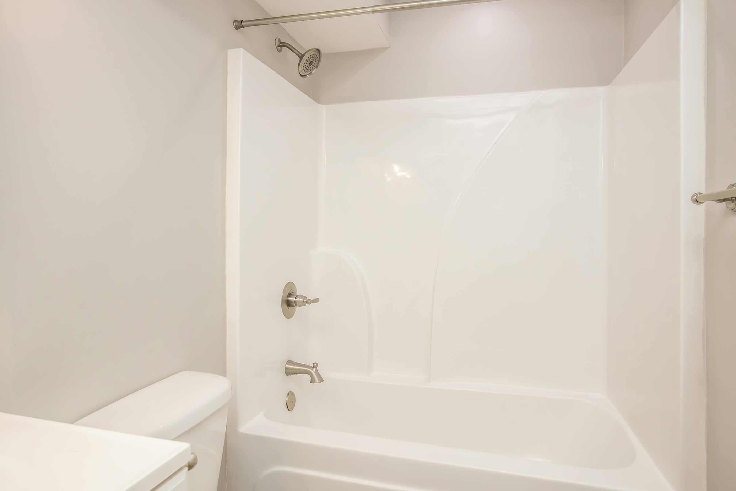 A clean, modern bathroom featuring a white bathtub and shower combination, a chrome showerhead, and a simple, sleek design. The walls are neutral-colored, enhancing the spacious and bright feel of the space. A toilet is visible to the left, adding to the bathroom's functionality.