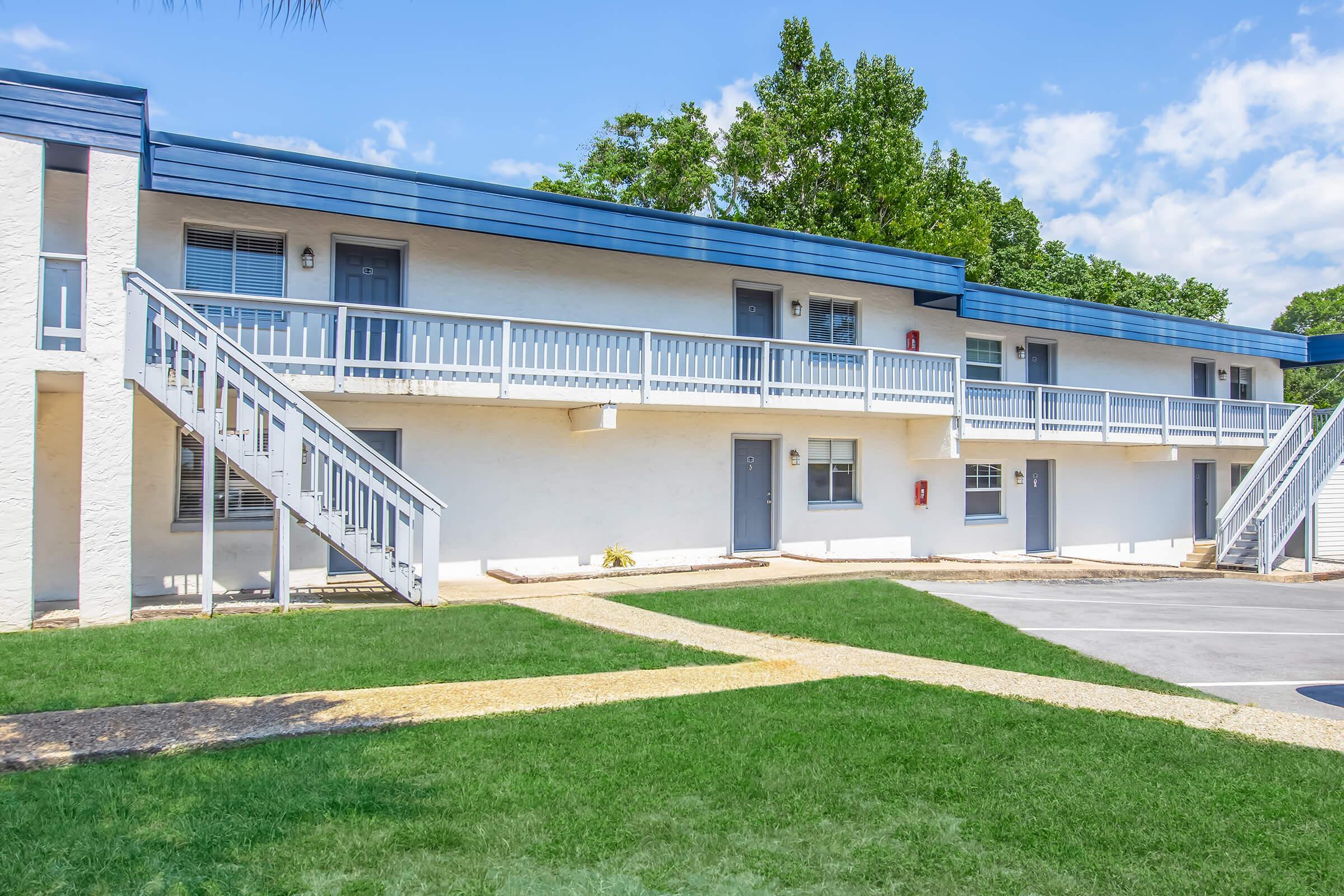 A two-story building with blue and white exterior features. It has multiple entrances with stairs leading up to the second floor. Green grass and a gravel pathway are visible in the foreground, with a bright blue sky and a few clouds in the background.