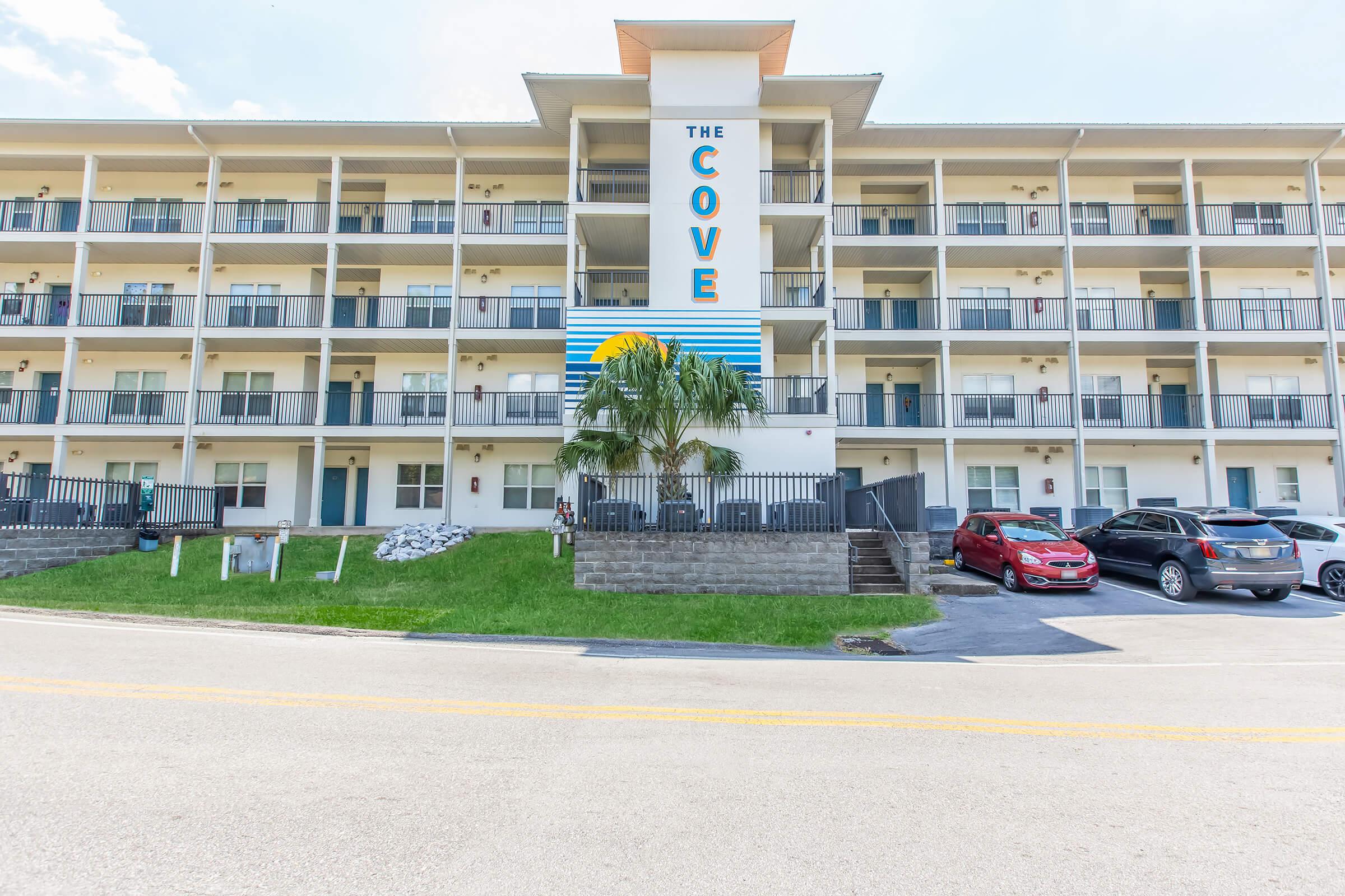 Exterior view of a three-story building named "The Cove," featuring balconies on each floor and palm trees in the front. Several parked cars are visible in the parking area, with a clear sky overhead and a well-maintained grassy area in front of the building.