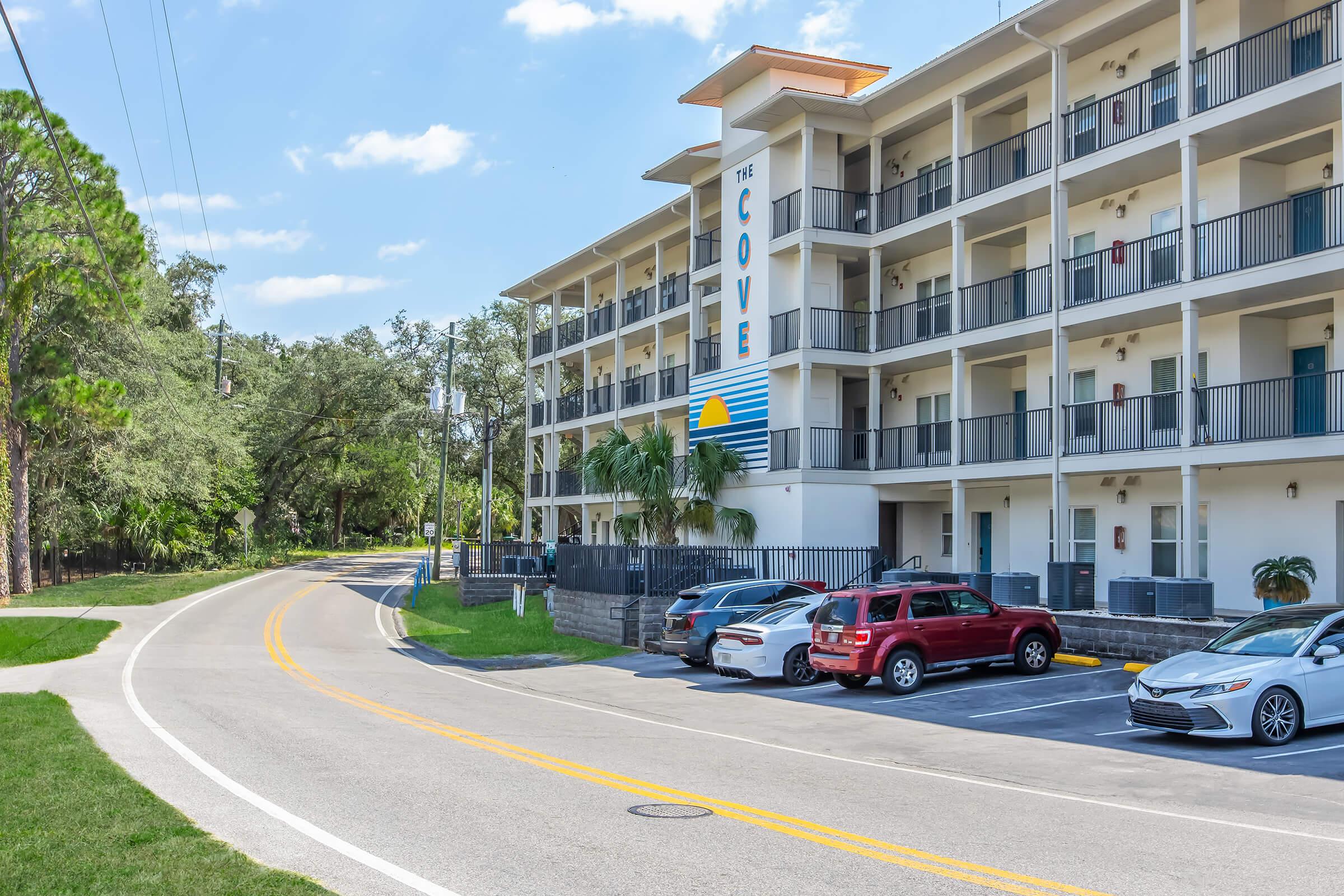 A view of a three-story building named "The Cove," featuring a modern architectural style with balconies. The road runs alongside the property, and several parked cars are visible. There are trees and greenery in the background, creating a pleasant environment. The sky is clear and blue.