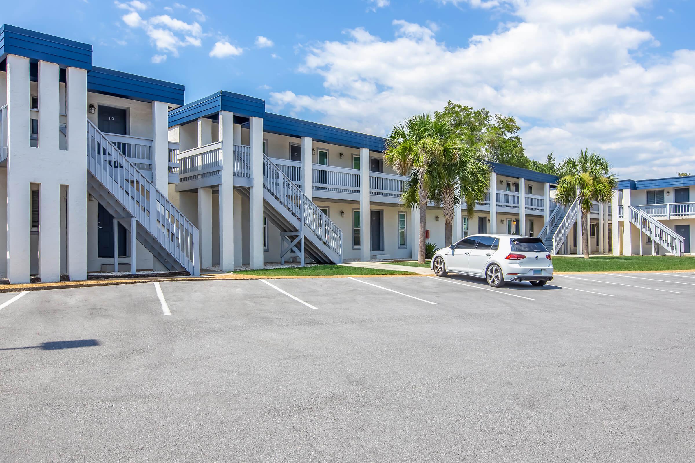 A two-story motel with prominent blue awnings and white walls. The building features staircases leading to the upper level, surrounded by green lawns and palm trees. A parked white SUV is visible in the foreground, with empty parking spaces lining the lot under a partly cloudy sky.