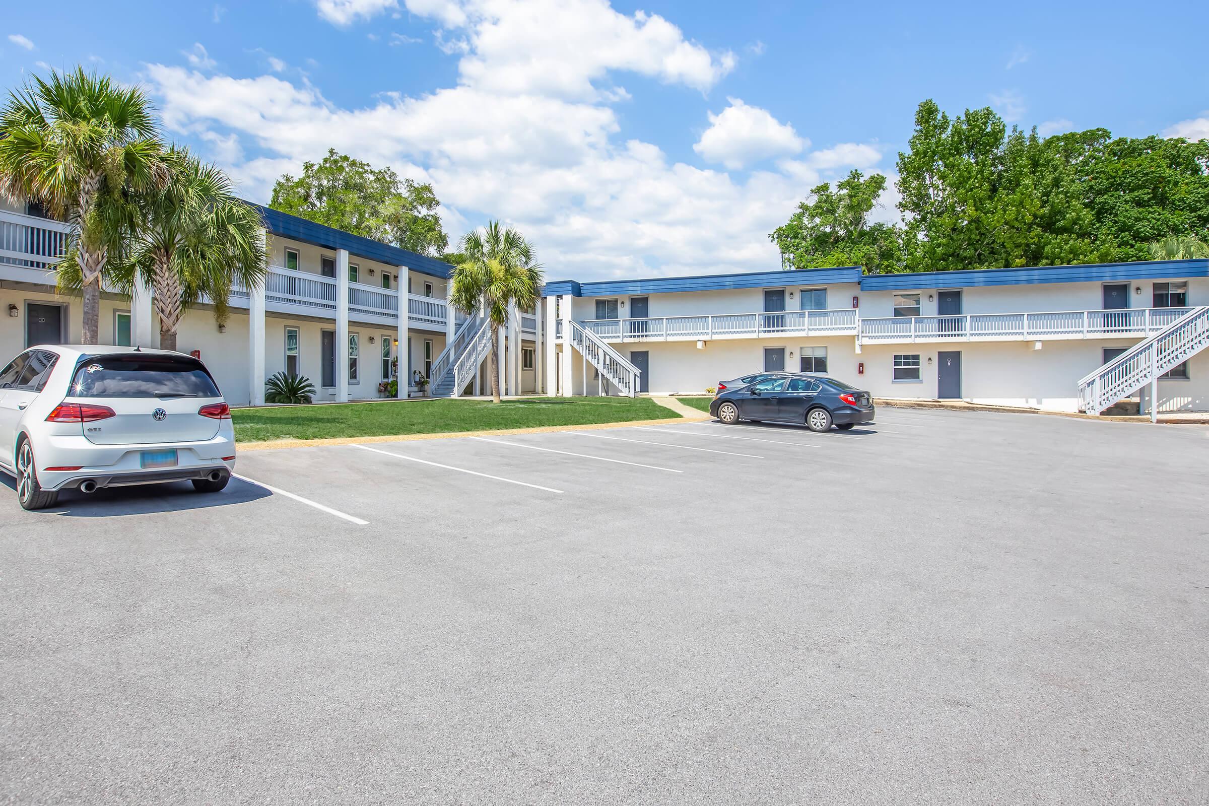 A view of a two-story motel with several rooms facing a parking lot. The exterior features blue roofs and white walls, surrounded by palm trees and greenery. Two parked cars are visible in the lot, and the sky is partly cloudy. The scene conveys a clean and inviting atmosphere.