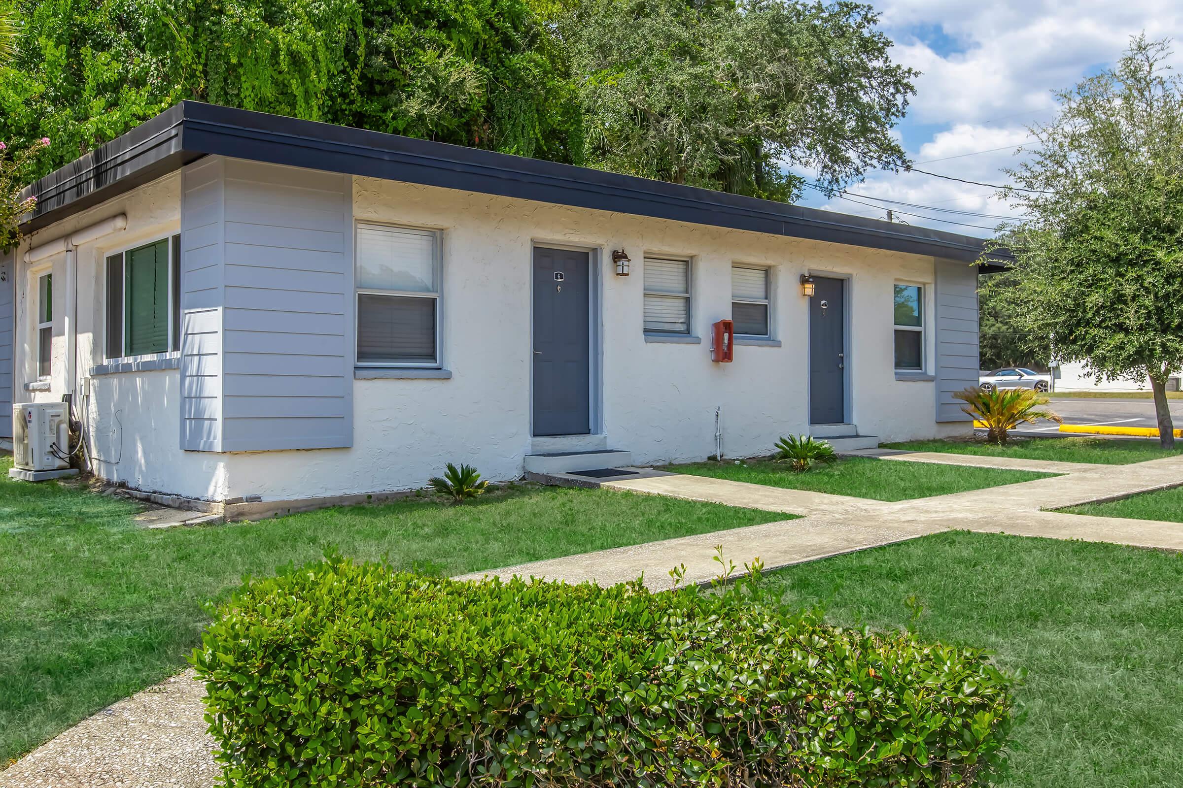A single-story residential building with a modern exterior, featuring light-colored walls and gray doors. There are two entrance doors visible, surrounded by green grass and small shrubs. The area is well-maintained, with a clear path leading to the doors and trees in the background under a partly cloudy sky.