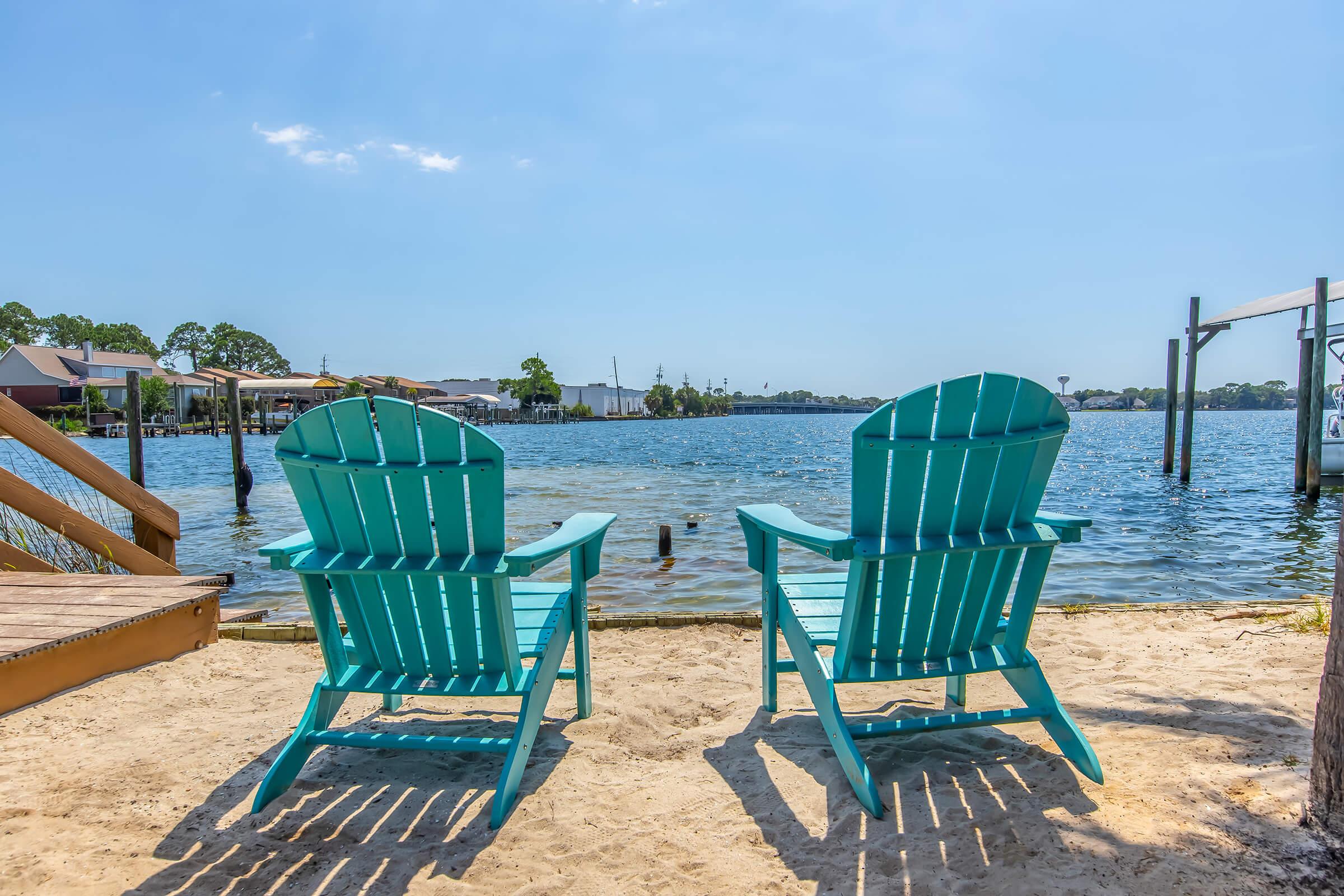 Two vibrant teal Adirondack chairs sit on sandy beach overlooking a calm body of water, with a clear blue sky above. In the distance, waterfront homes and boats are visible, creating a tranquil and inviting atmosphere perfect for relaxation.