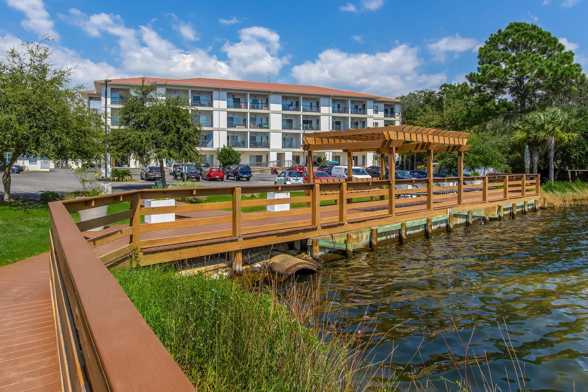 A scenic view of a lakeside wooden deck with a pergola, surrounded by lush greenery. In the background, a multi-story building is visible, with several parked cars nearby. The sky is partly cloudy, creating a pleasant atmosphere by the water.