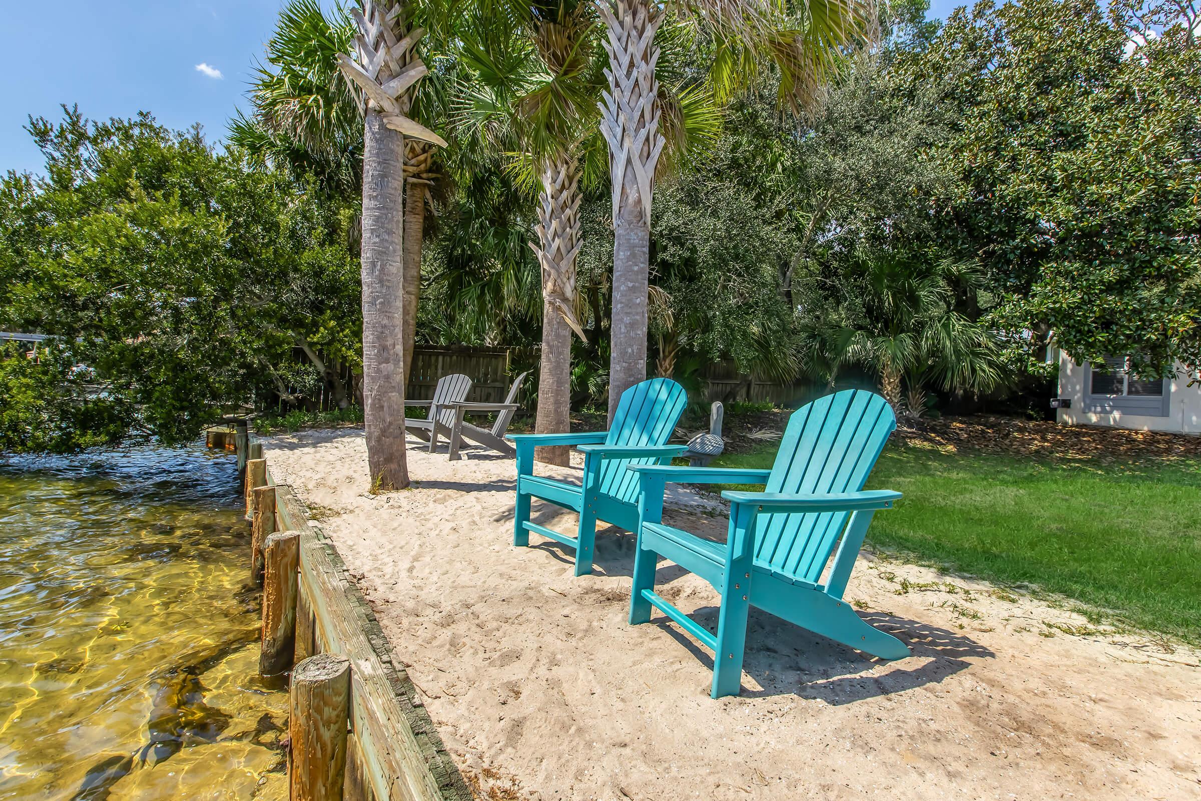Two brightly colored turquoise adirondack chairs sit on a sandy area near a calm body of water, surrounded by lush greenery and palm trees. The scene captures a serene outdoor space perfect for relaxation, with clear blue skies overhead and gentle reflections on the water's surface.