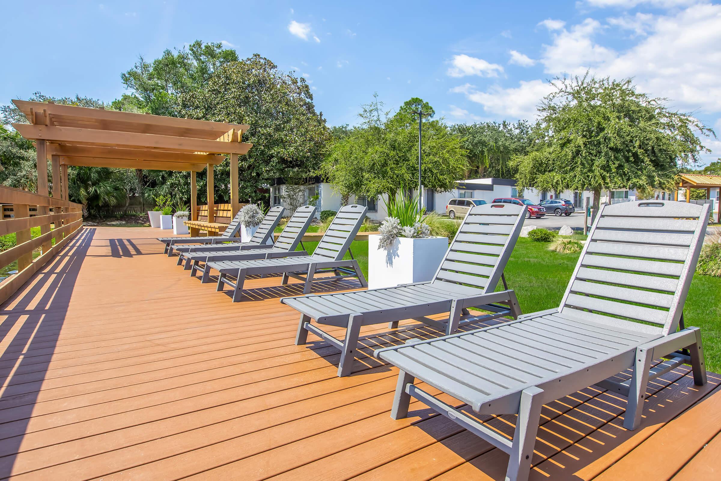 A sunny outdoor area featuring several gray lounge chairs arranged on a wooden deck. In the background, there are green trees, a pergola, and a well-maintained lawn. The scene conveys a relaxing atmosphere ideal for leisure and enjoyment.