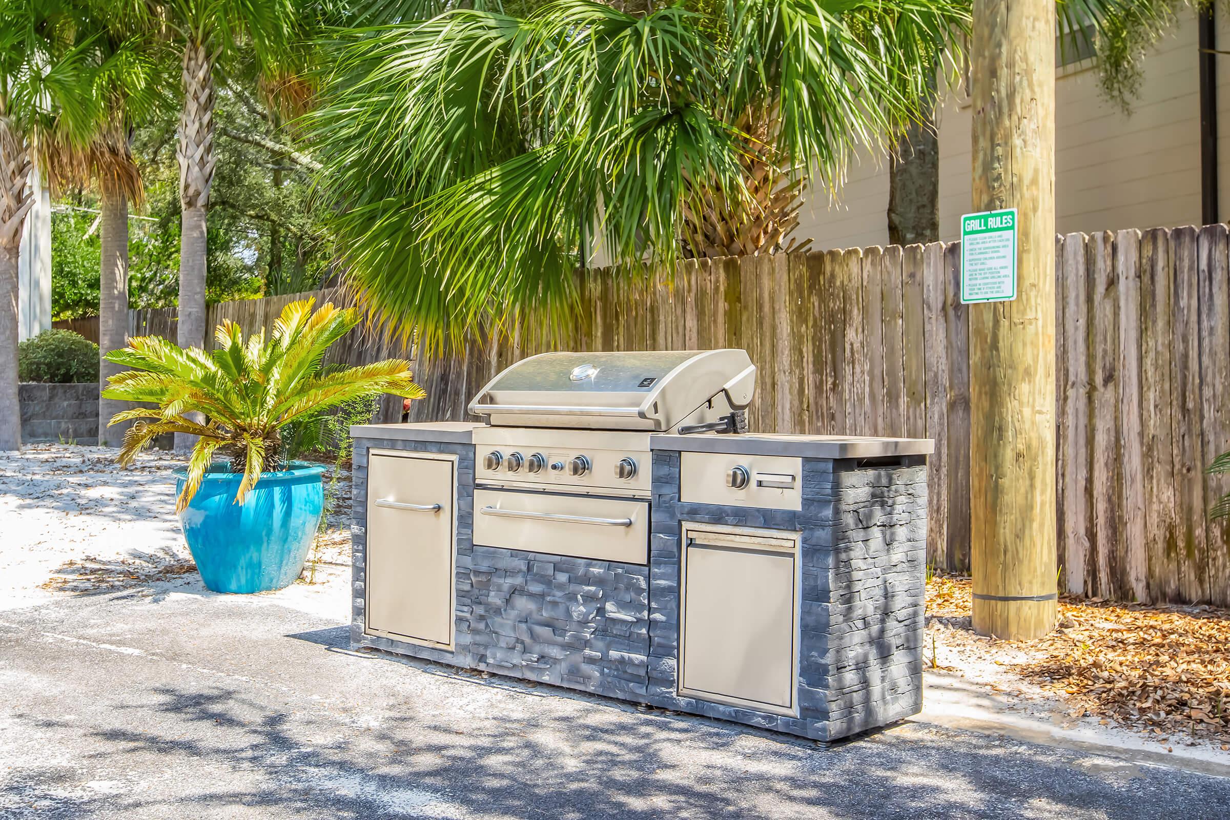 An outdoor grilling station featuring a stainless steel gas grill, stone countertops, and storage cabinets. Surrounded by palm trees and decorative plants, the setup is placed on a paved surface next to a wooden fence and a utility pole.